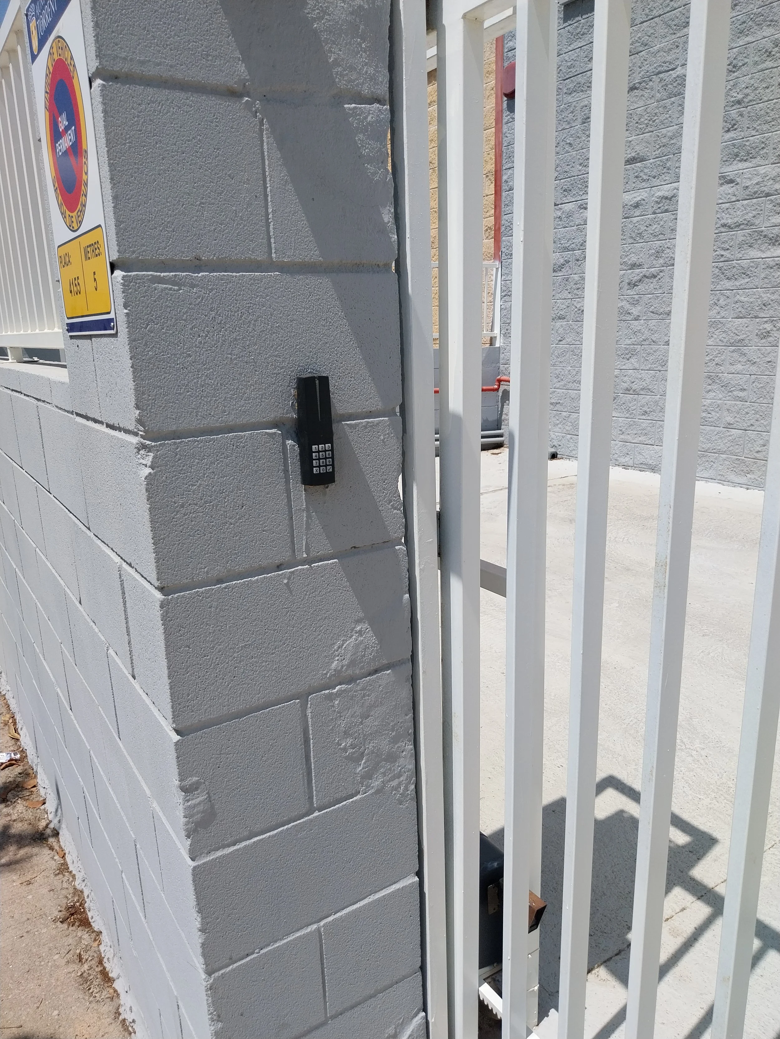 Muro de bloques de concreto pintados de gris y pórtico de reja blanca decorativa, con un código de seguridad en un muro y señal de no estacionar.