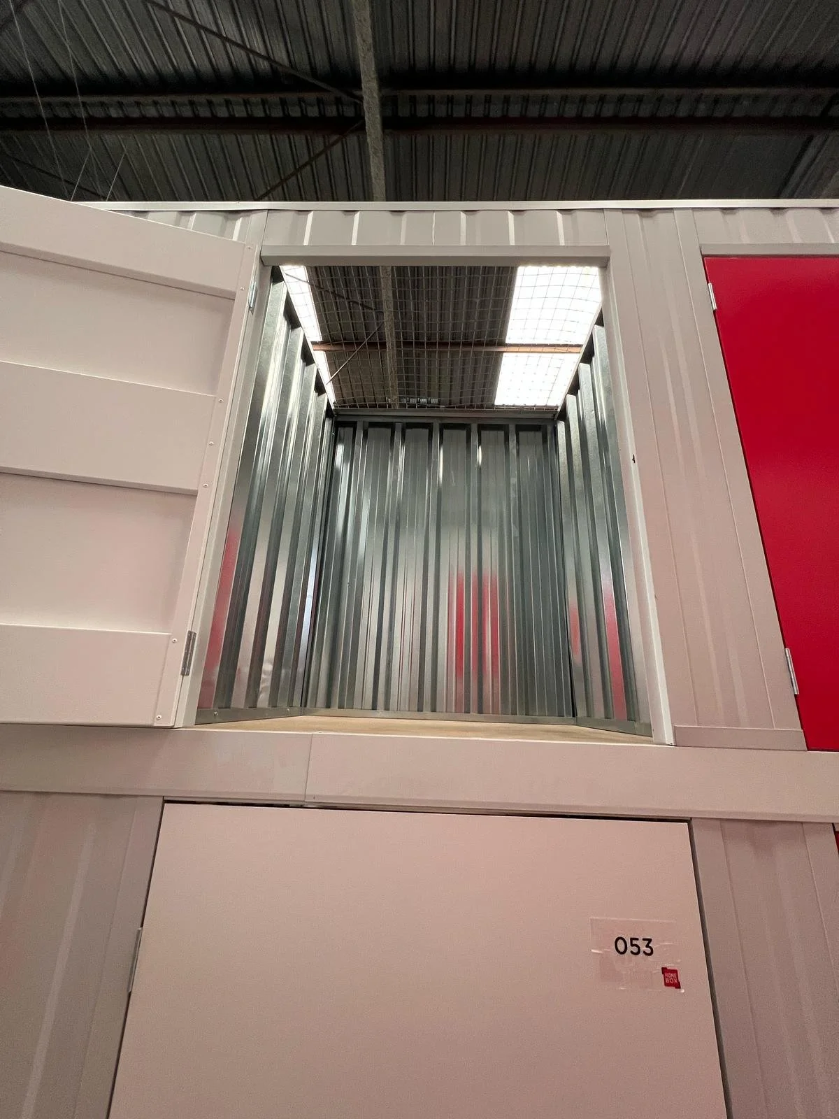 Interior de una tienda con un espacio para almacenamiento o exhibición, con paredes de metal y un techo de láminas de metal, en el centro un espacio vacío y en la parte inferior una puerta blanca con la etiqueta de número 053.