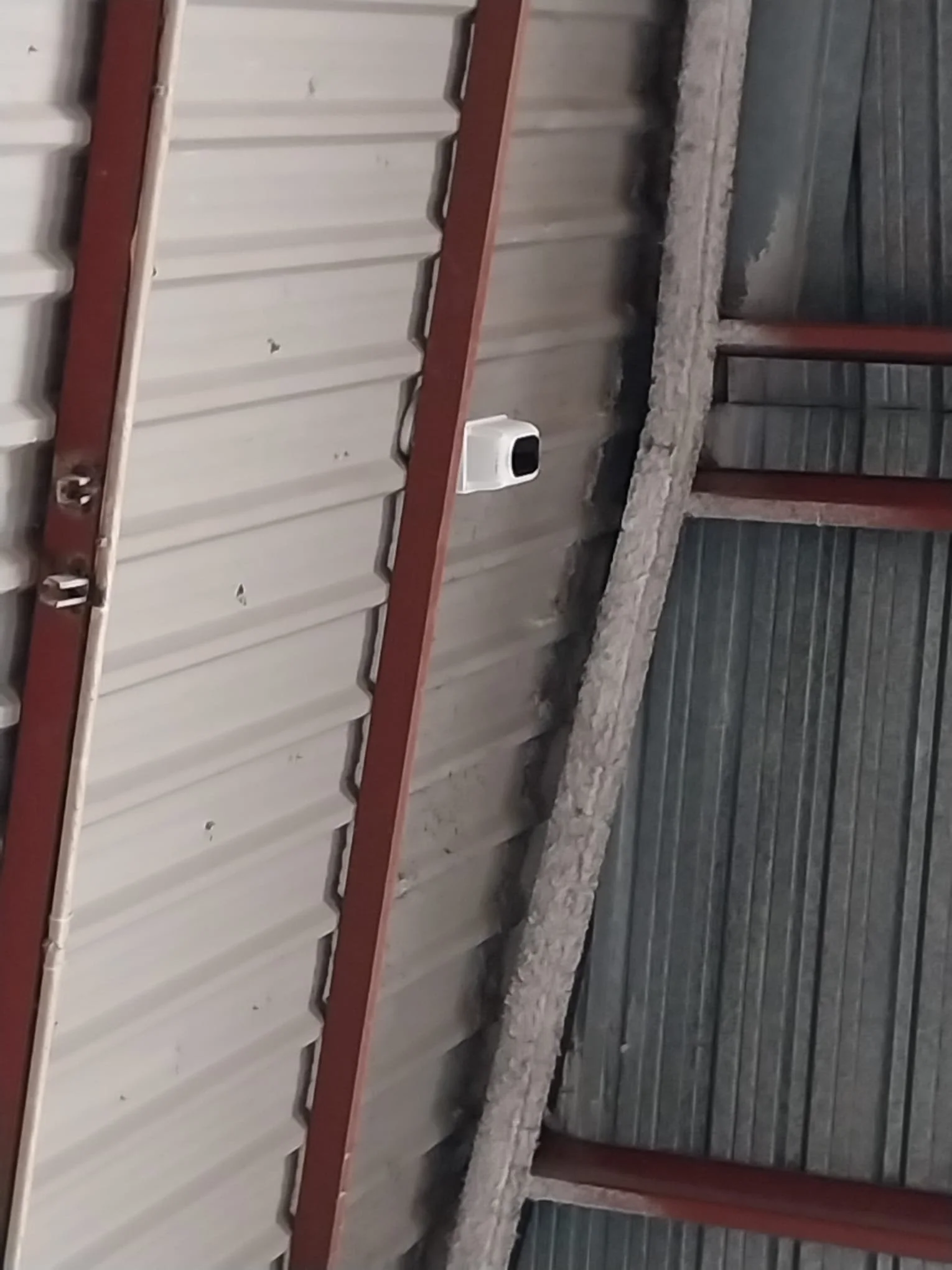 Una pared de láminas metálicas de color gris con estructura de madera y una cámara de vigilancia blanca instalada en la pared.