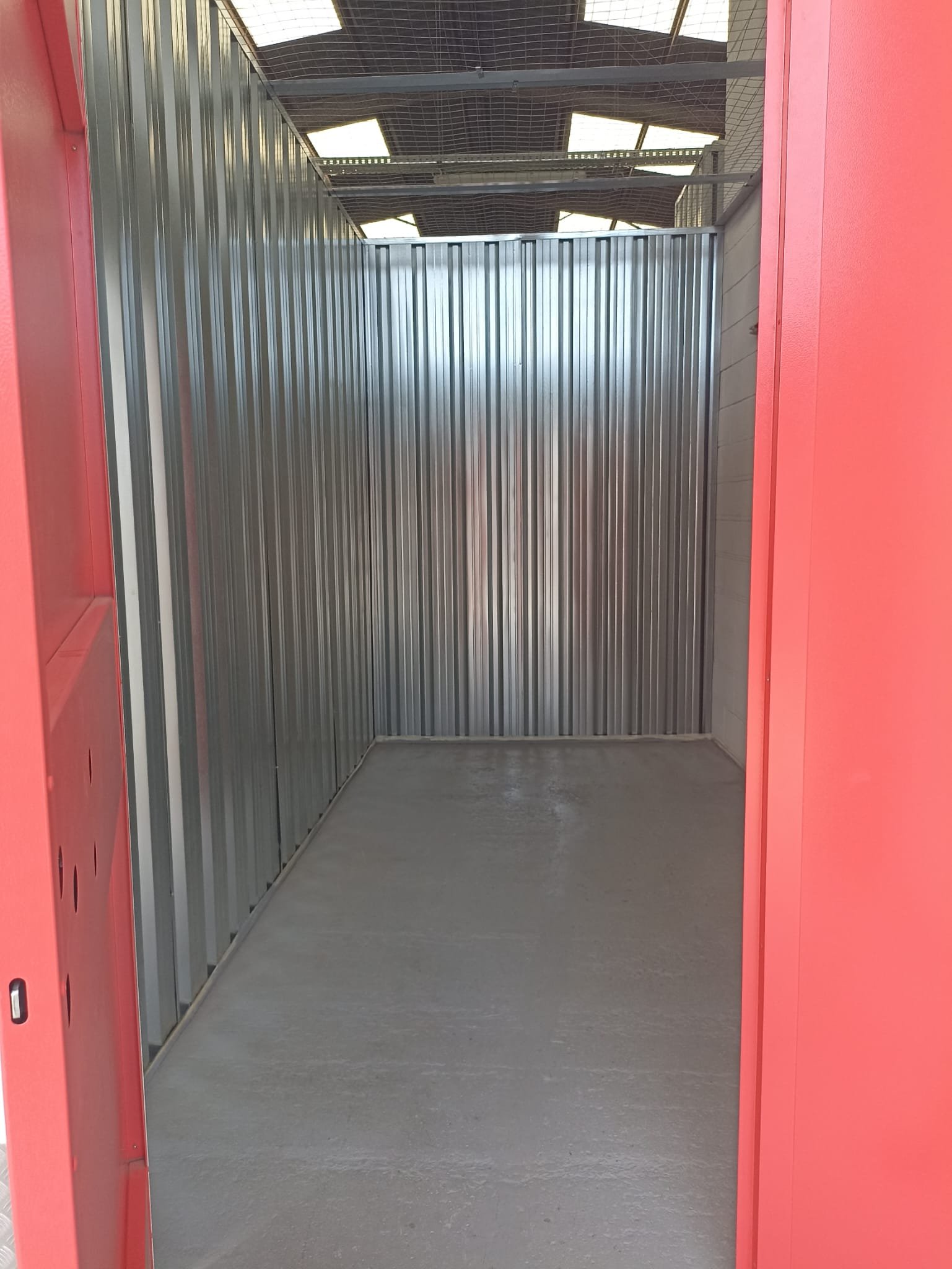 Una entrada de ascensor vacía con paredes rosadas a los lados y puertas abiertas. La cabina tiene paredes de metal pulido y un piso de concreto. En el techo, se ven paneles de cristal parcialmente cubiertos con una estructura metálica.