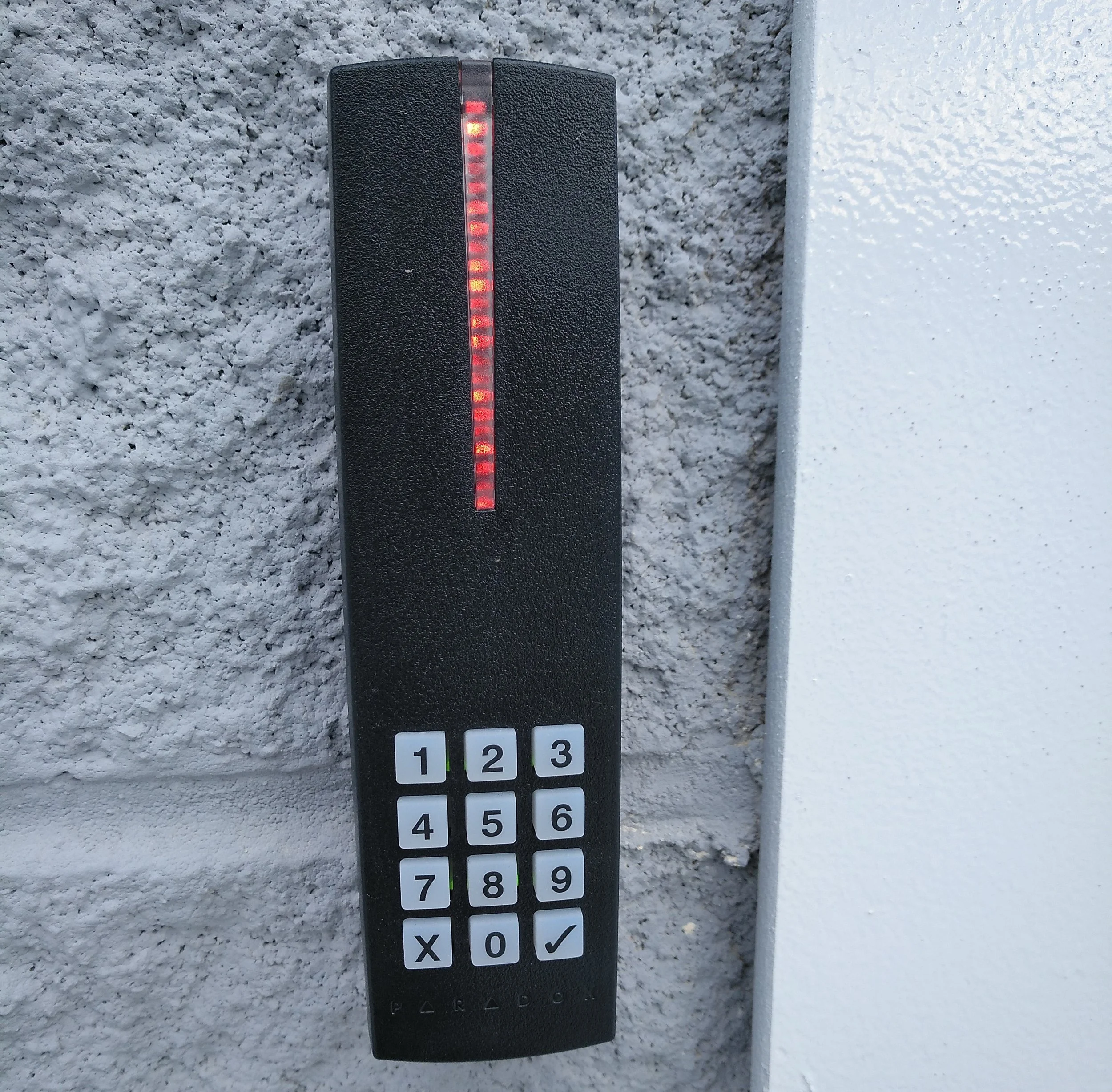 Dispositivo electrónico negro con una línea vertical de luces rojas y amarillas, y una fila de botones numerados del 1 al 9 y otros botones con símbolos, colocado sobre una pared de superficie rugosa gris.