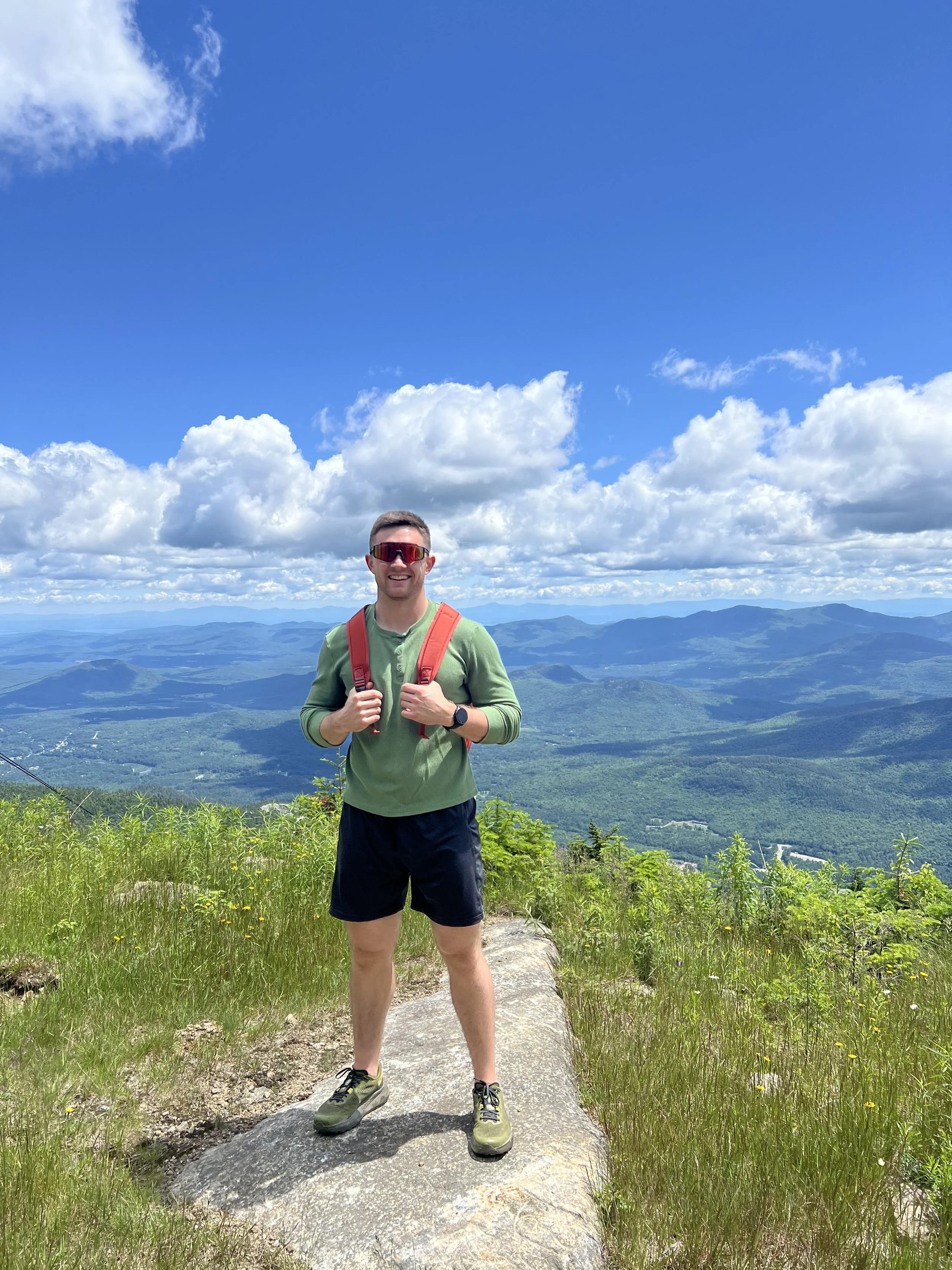 A man in outdoor hiking gear standing on a large rock with a scenic mountain and valley view behind him, under a partly cloudy sky.