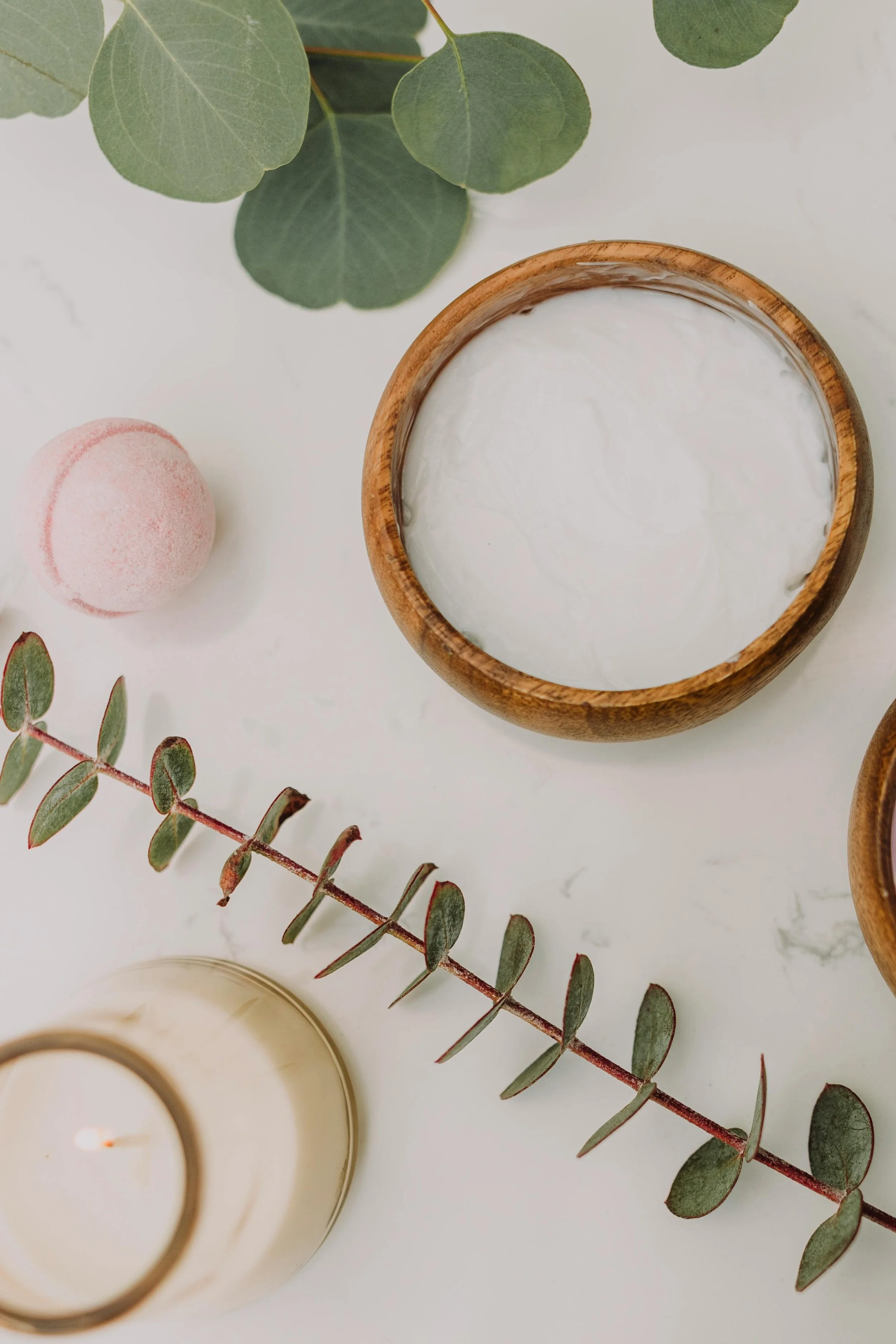 A top-down view of a wooden bowl filled with white cream, a pink bath bomb, a white votive candle, and green eucalyptus leaves on a white surface.