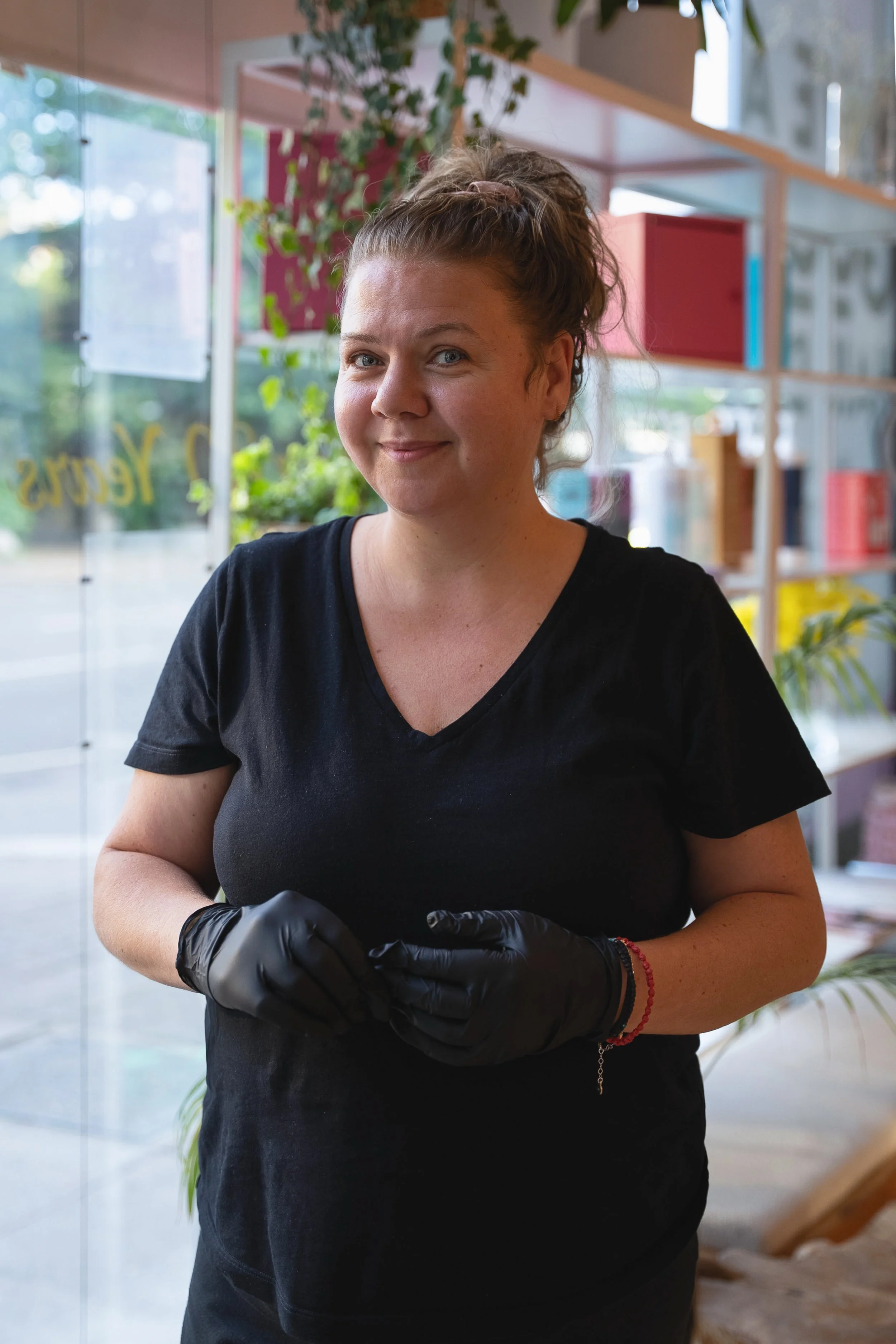 A woman wearing a black shirt and black gloves standing inside a modern space with glass walls, shelves filled with colorful objects, and green plants in the background.