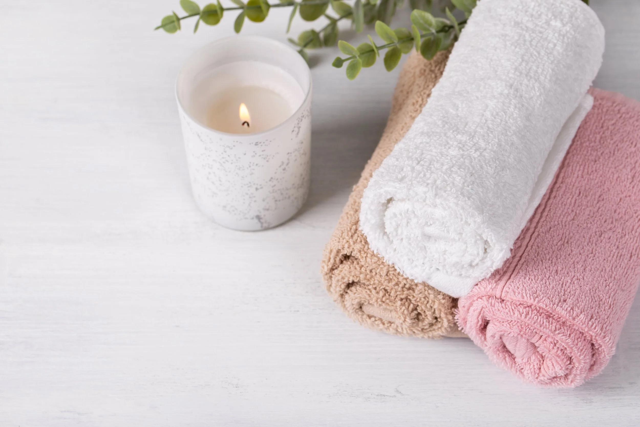 Stacked towels in white, beige, pink, and cream, with a lit candle and green foliage on a white surface.