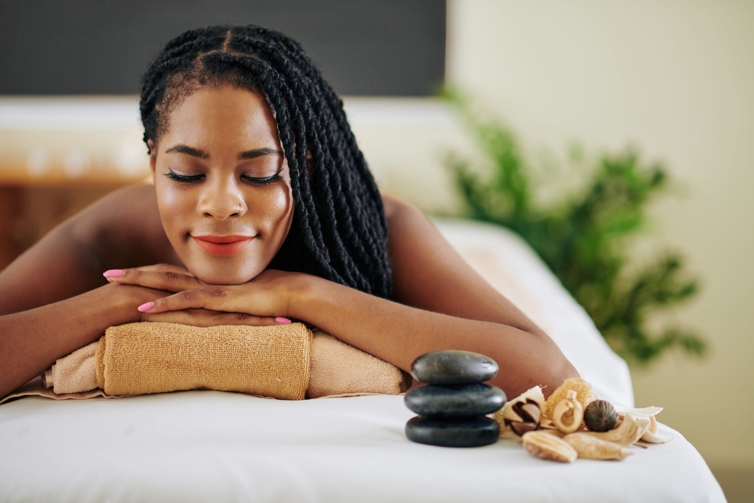 A woman with braided hair resting her head on her folded arms during a massage, with spa stones and shells on a white towel, in a relaxing spa setting.