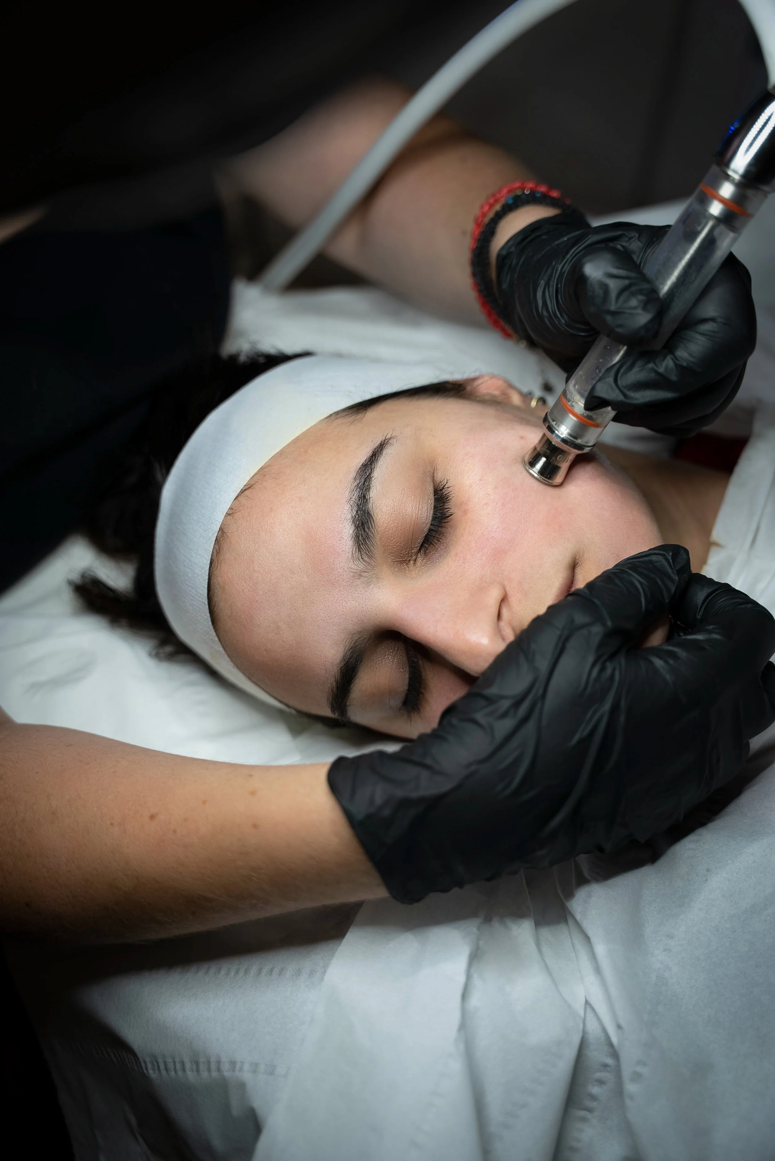 A woman receives a hydrafacial treatment with a handheld device by a professional wearing black gloves, lying on a medical bed with a white headband and a white gown.