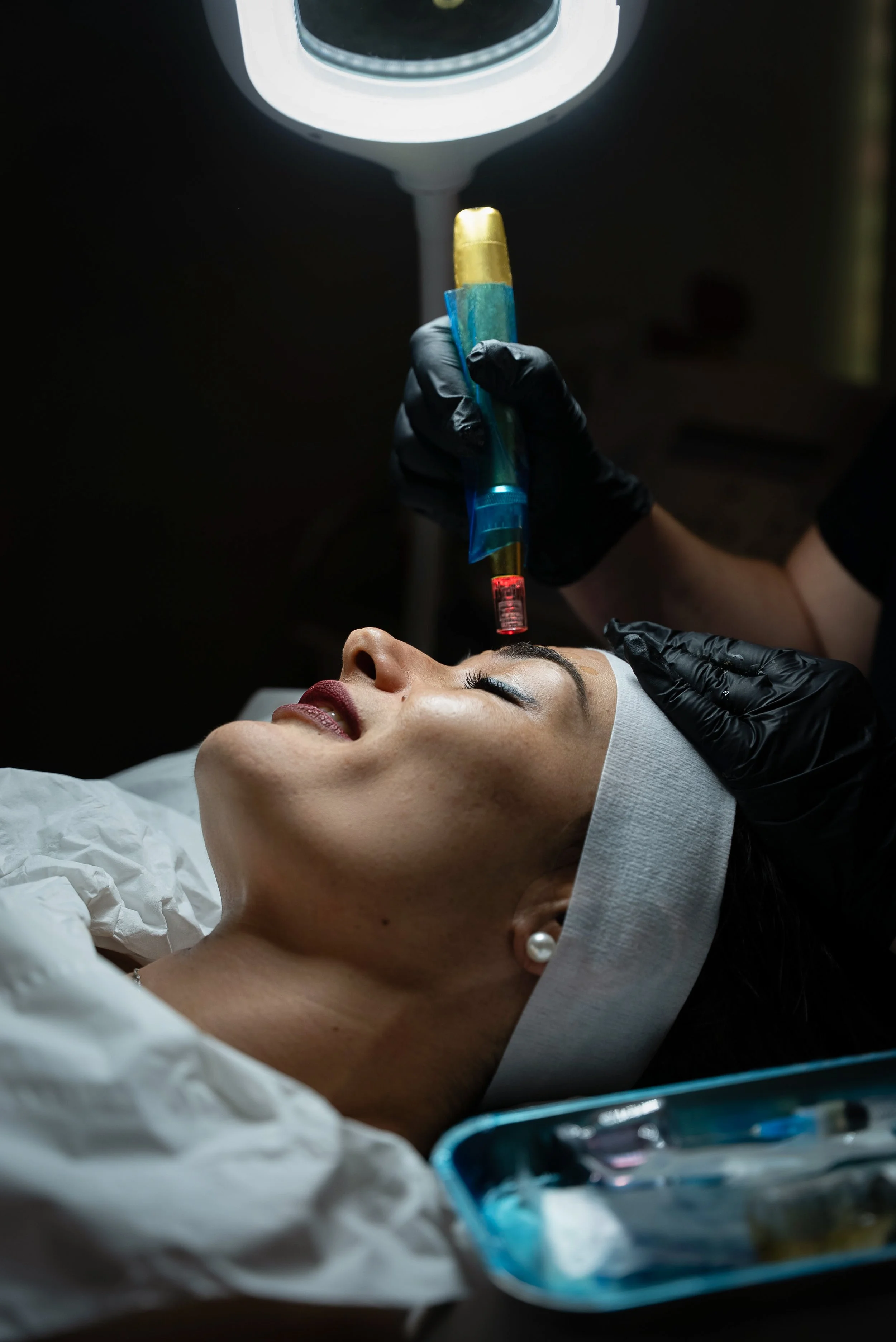A woman receives a cosmetic injection or treatment in a clinical setting, lying down with her eyes closed, while a healthcare professional in black gloves administers the procedure using a syringe. The scene is dimly lit, emphasizing the medical procedure.
