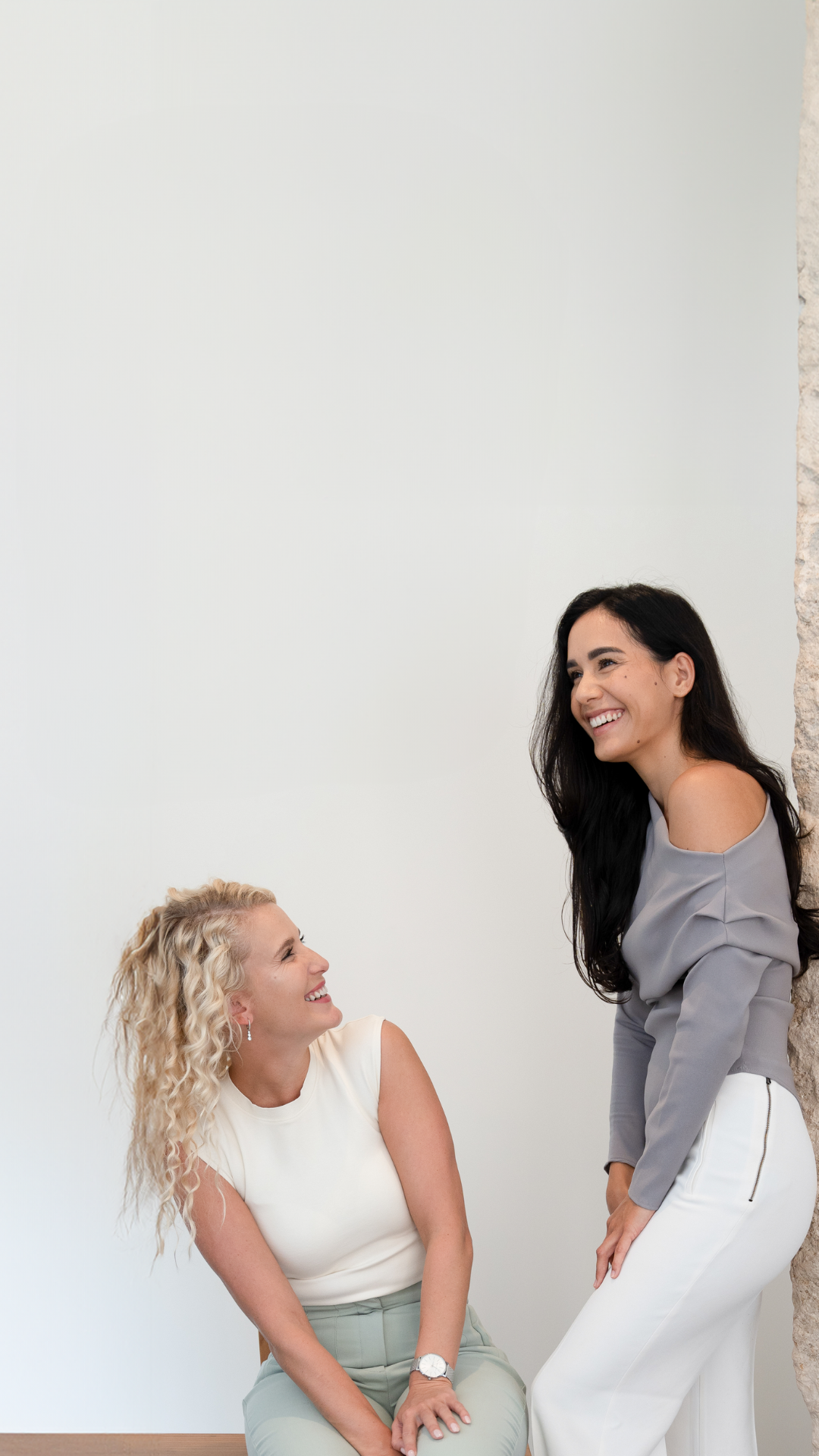 Two women smiling and laughing, one with curly blonde hair sitting down and the other with long black hair standing, against a white wall with a stone column.