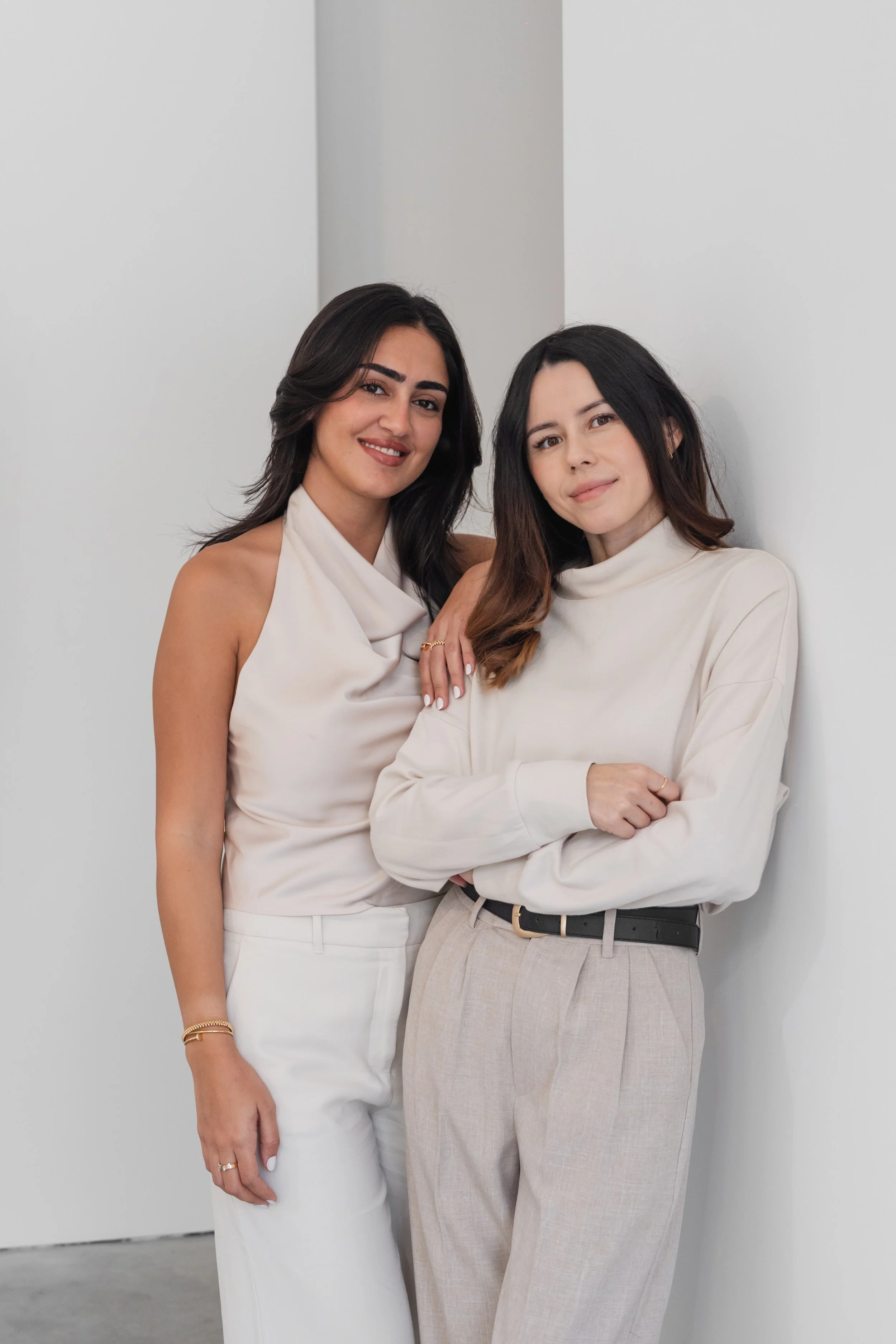 Two women standing close together against a white wall, smiling at the camera. One woman has dark hair, wearing a satin sleeveless top and white pants. The other woman has light brown hair, wearing a cream-colored turtleneck and beige pants.