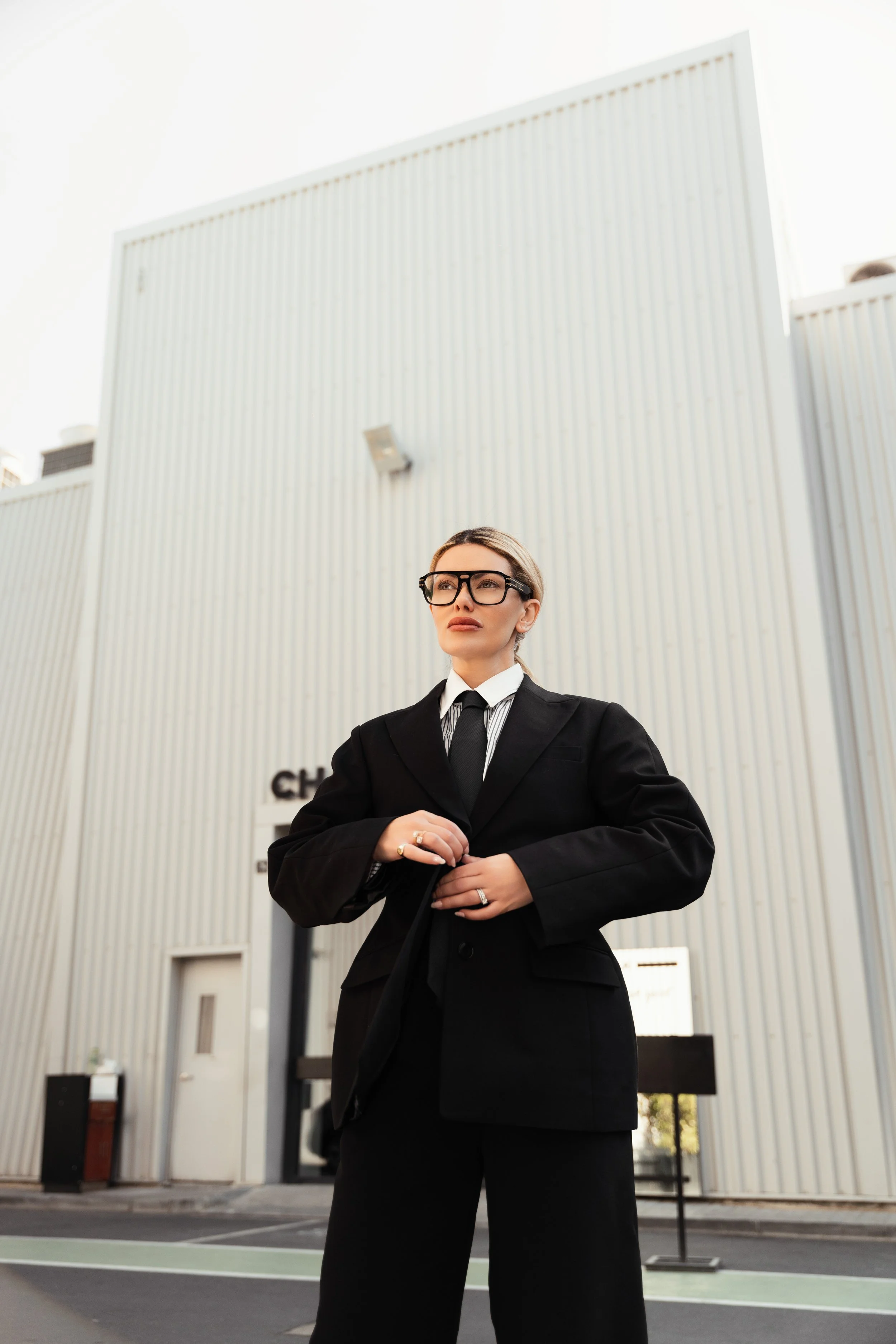 Businesswoman wearing glasses and a black suit standing outside a modern building with vertical corrugated metal walls.