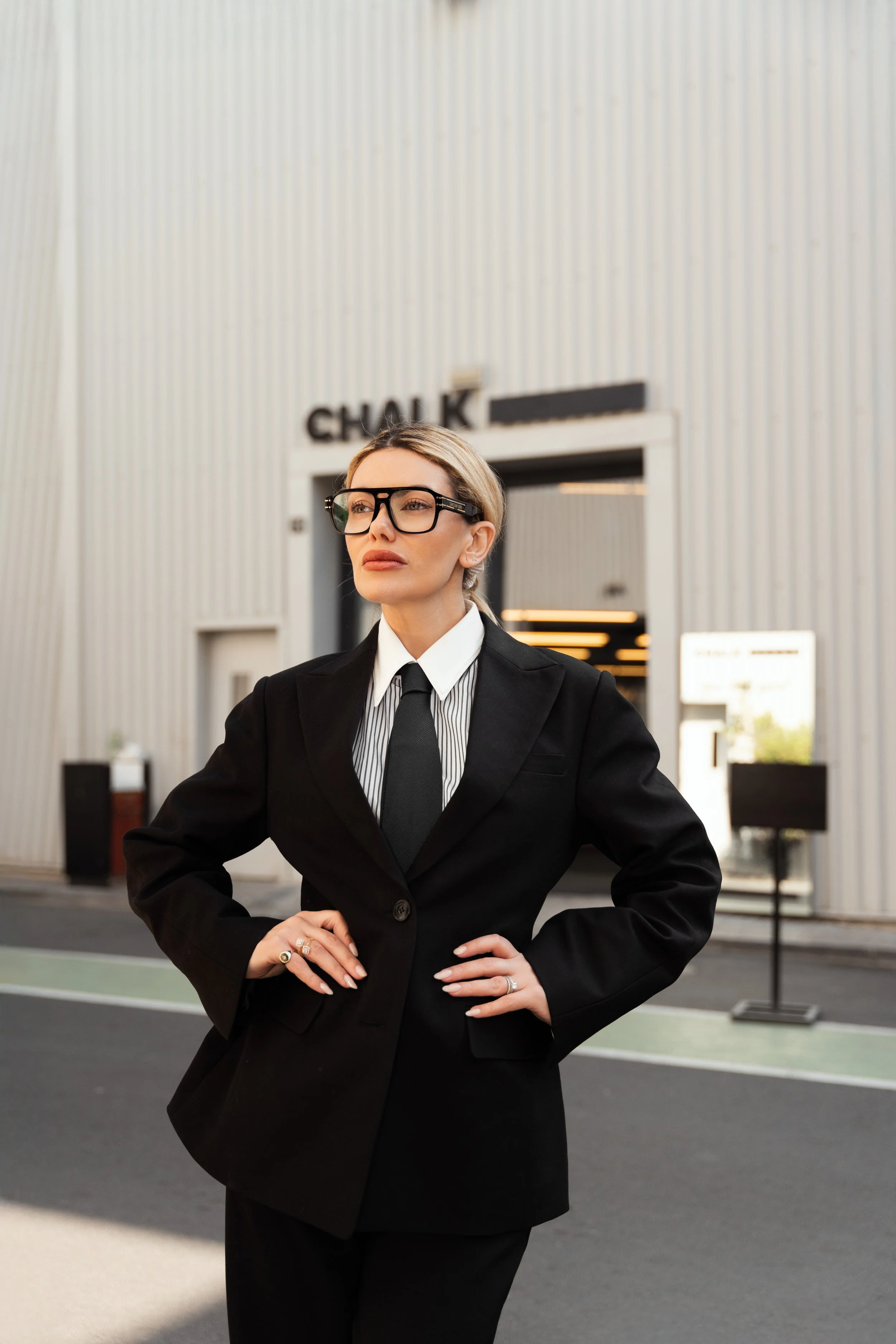 A woman in a black suit with glasses standing outside a modern building with vertical paneling and the sign 'CHALK' above the entrance.