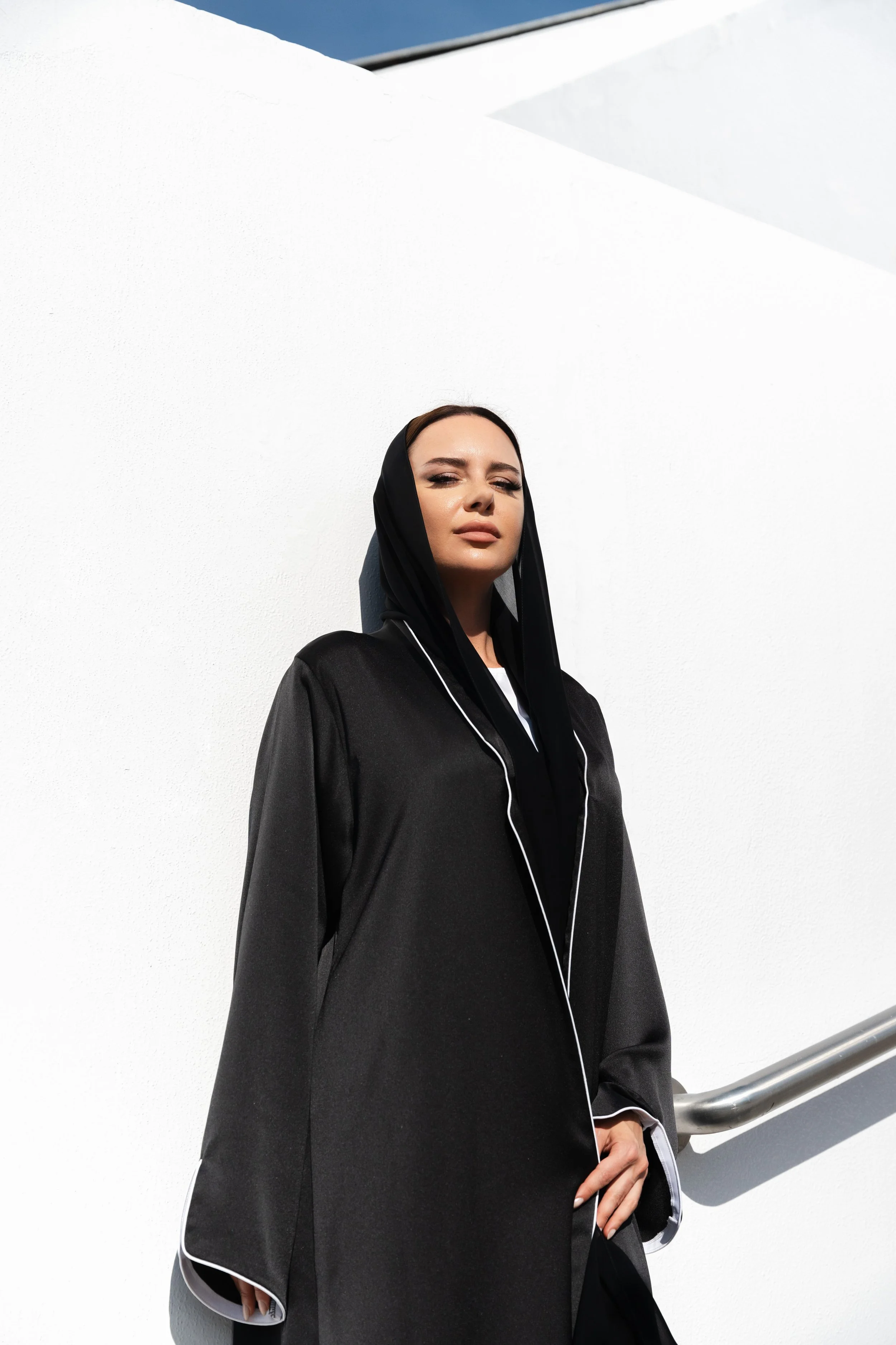 A woman standing against a white wall, wearing a black hooded garment with white piping, looking confidently at the camera.