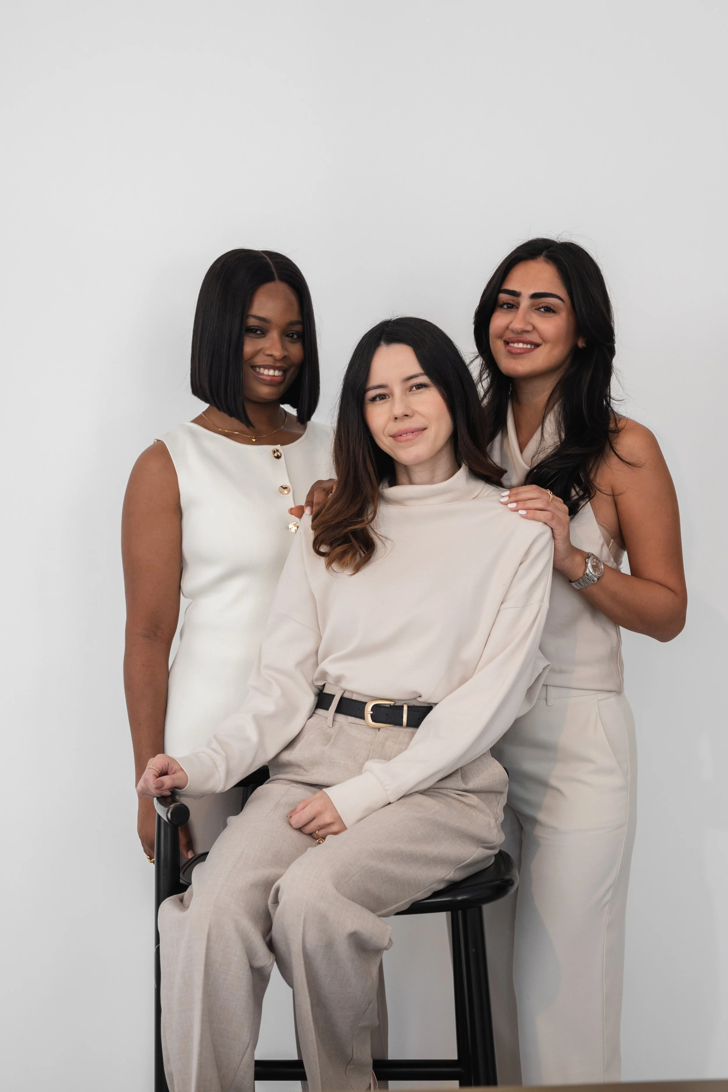 Three women posing together against a plain white background. The woman in the middle is seated on a stool, wearing a cream-colored turtleneck and beige pants, with dark wavy hair and a soft smile. The woman on the left has dark skin, straight bob ha