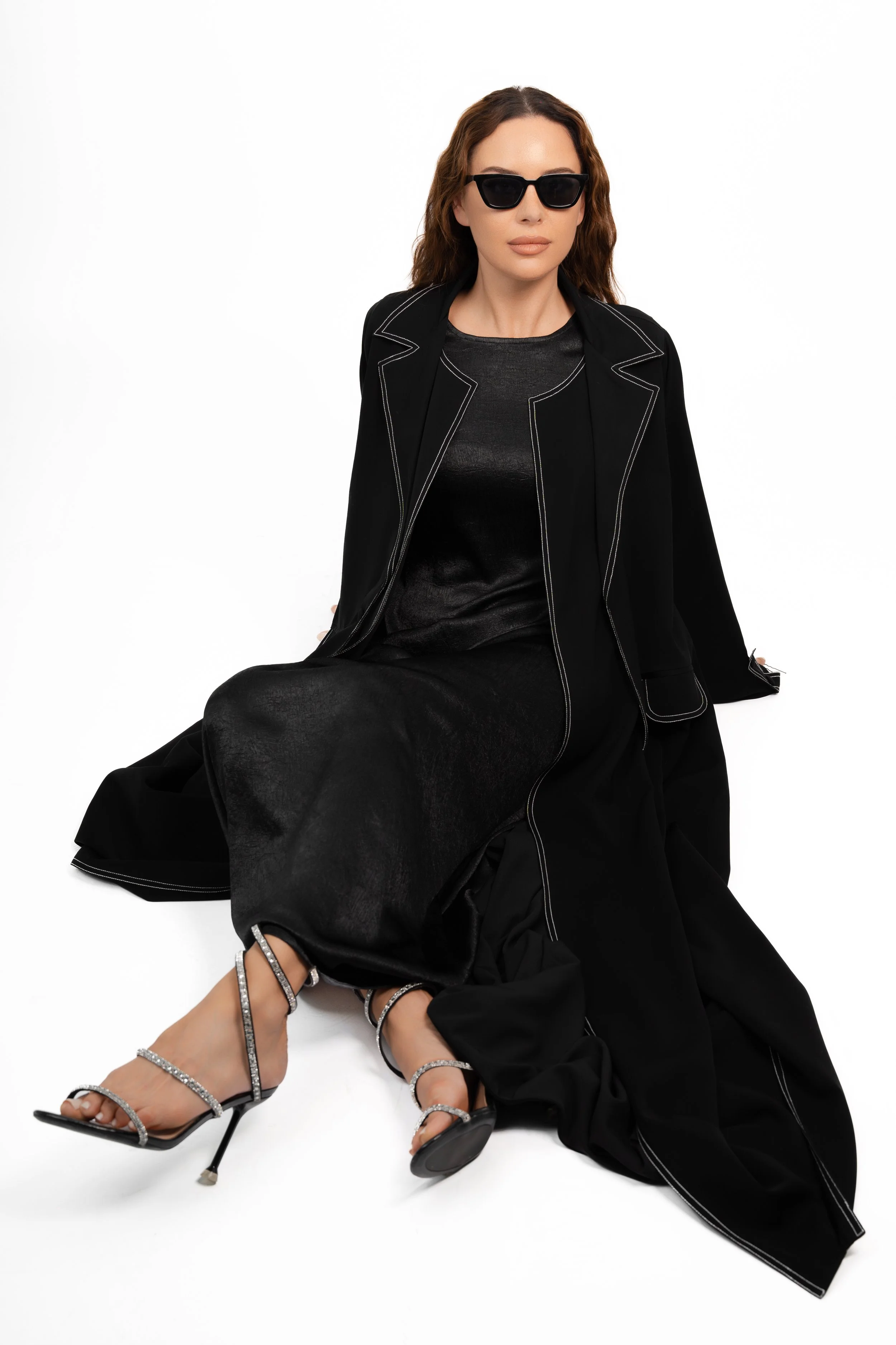 A woman sitting on the floor wearing black sunglasses, a black jacket, a black dress, and silver high-heeled sandals with rhinestones.