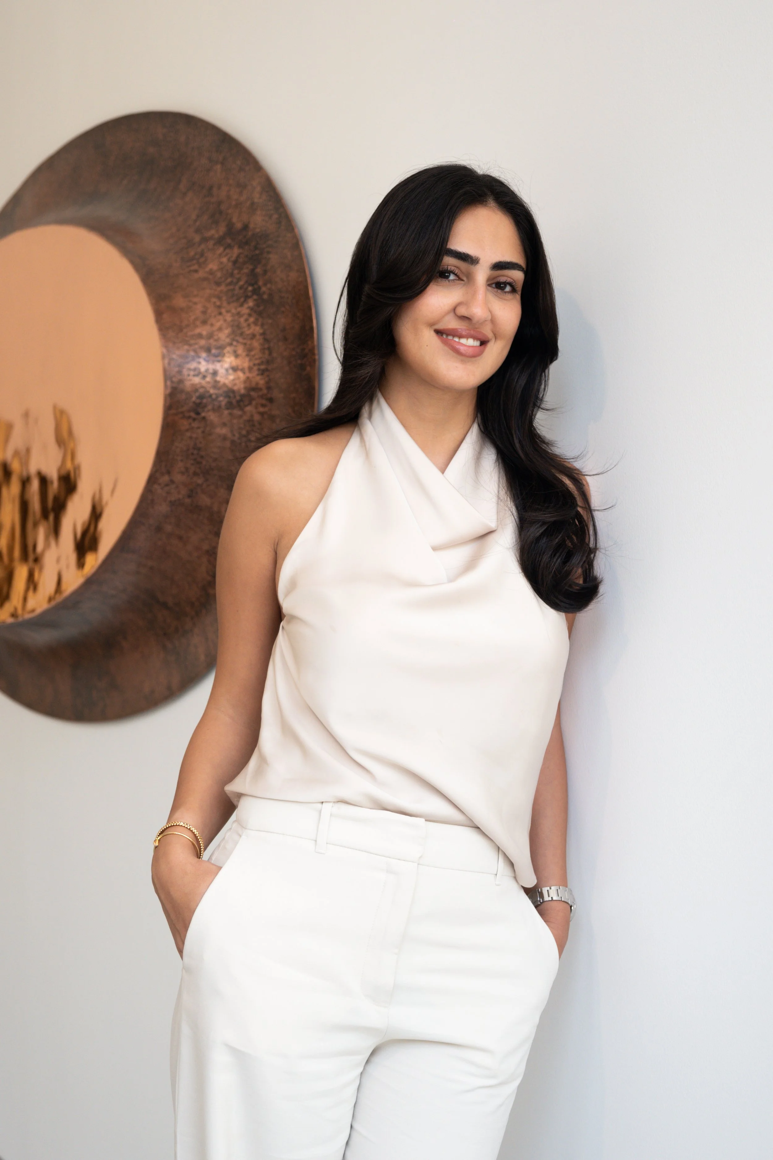 A woman with long dark hair, dressed in a sleeveless white top and white pants, standing against a white wall with a circular piece of artwork behind her.