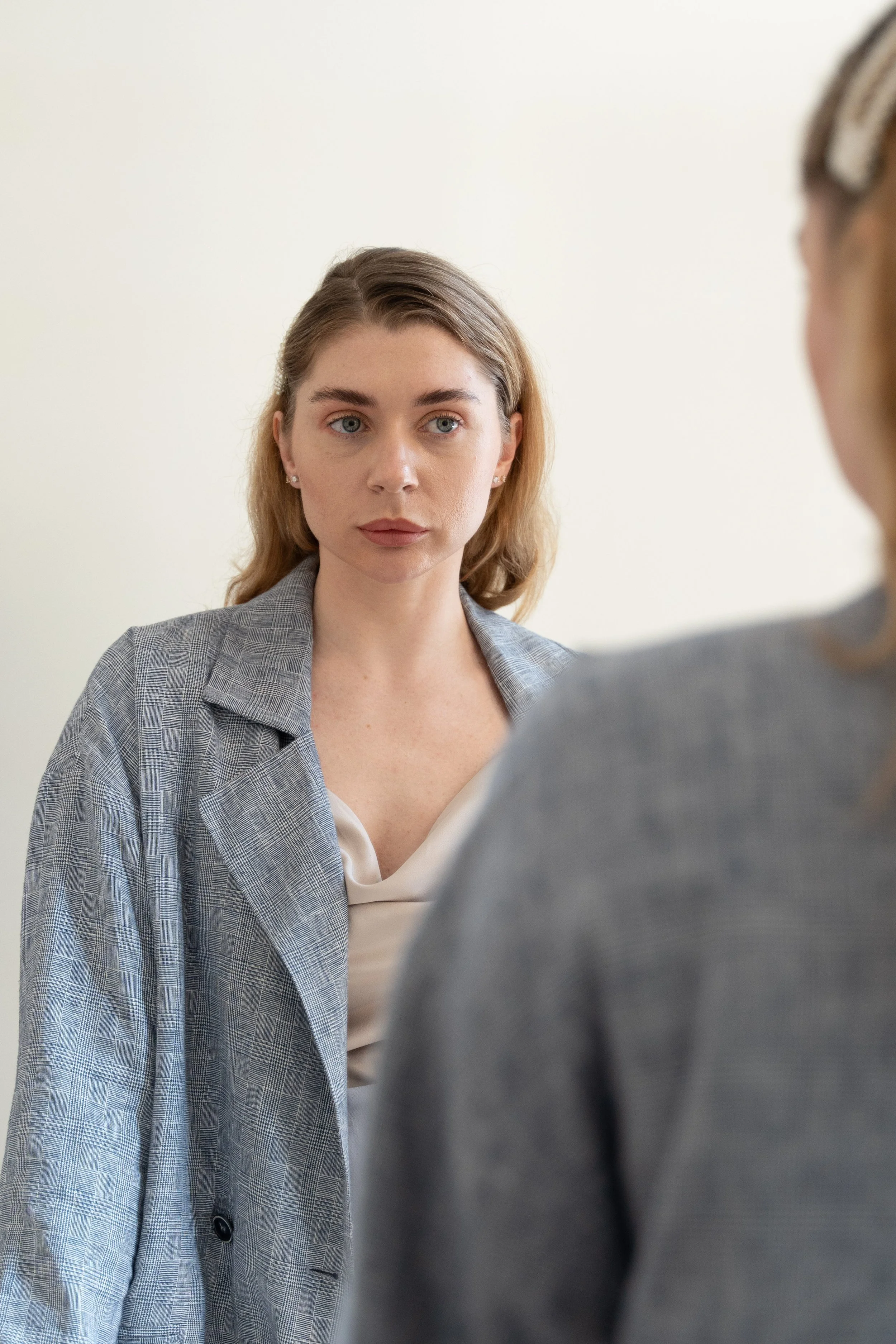 A woman with light brown hair and blue eyes looking at another woman with blurred features in a gray blazer.
