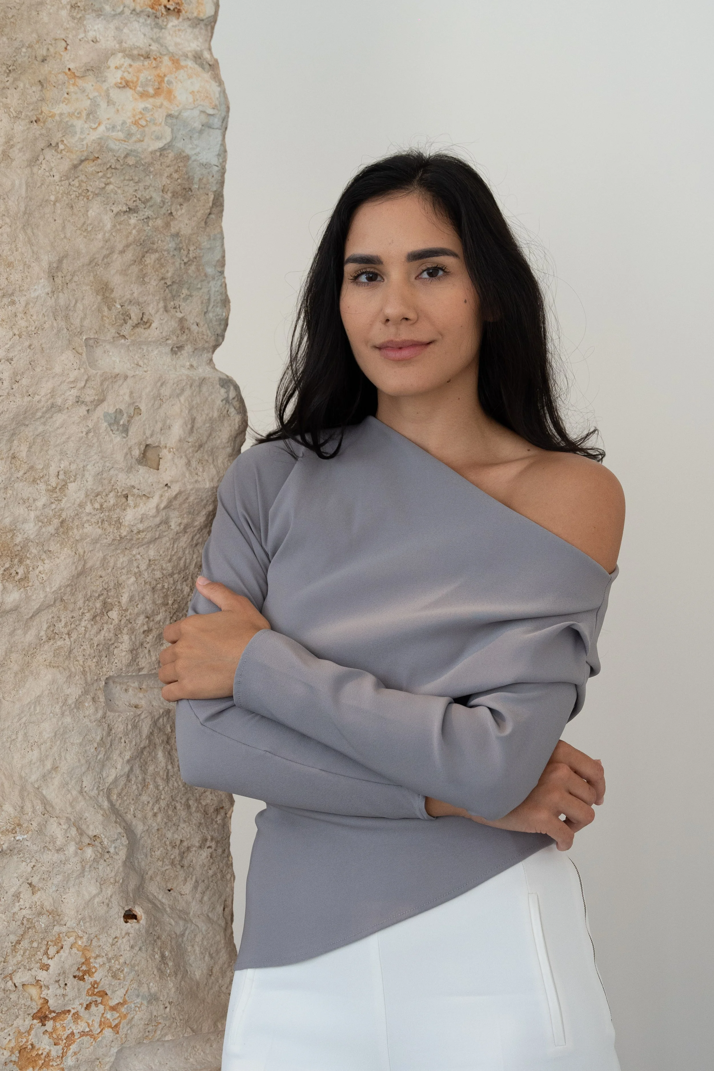 A woman with long dark hair, wearing a off-the-shoulder light gray top and white skirt, standing next to a textured stone wall with a neutral background.