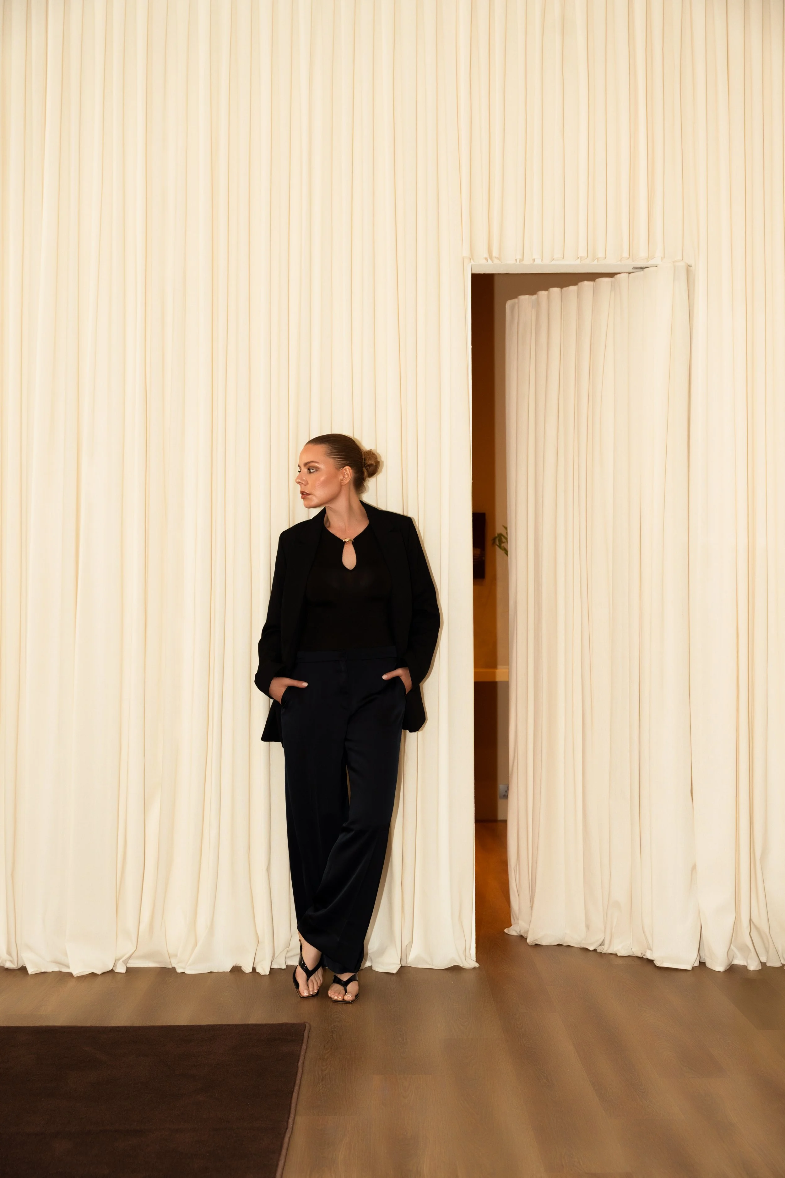 Woman in black suit standing against beige curtains, with hands in pockets, looking to the side.