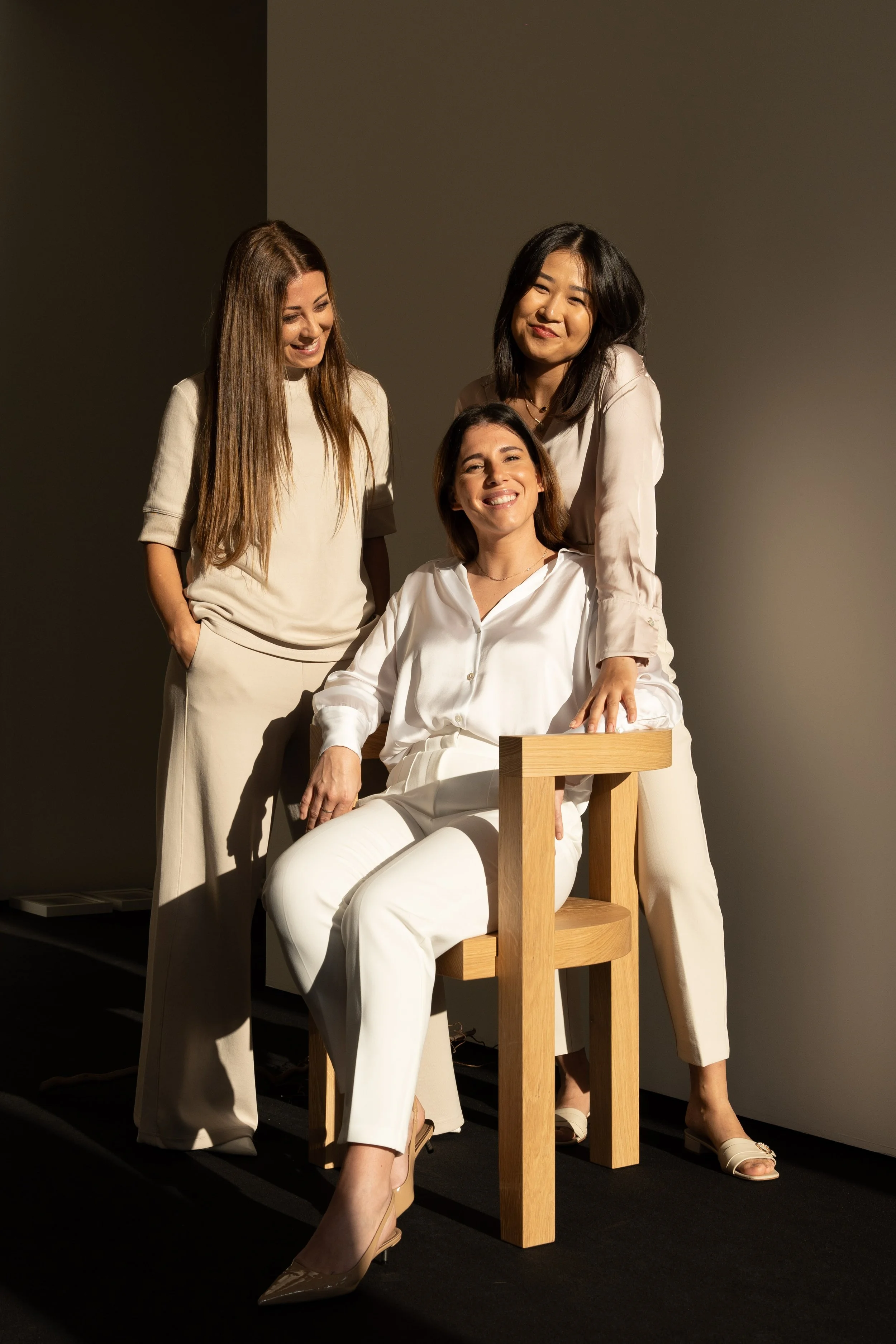 Three women smiling and posing in a photography studio, with one sitting on a wooden chair and two standing beside her, dressed in white and cream-colored outfits.