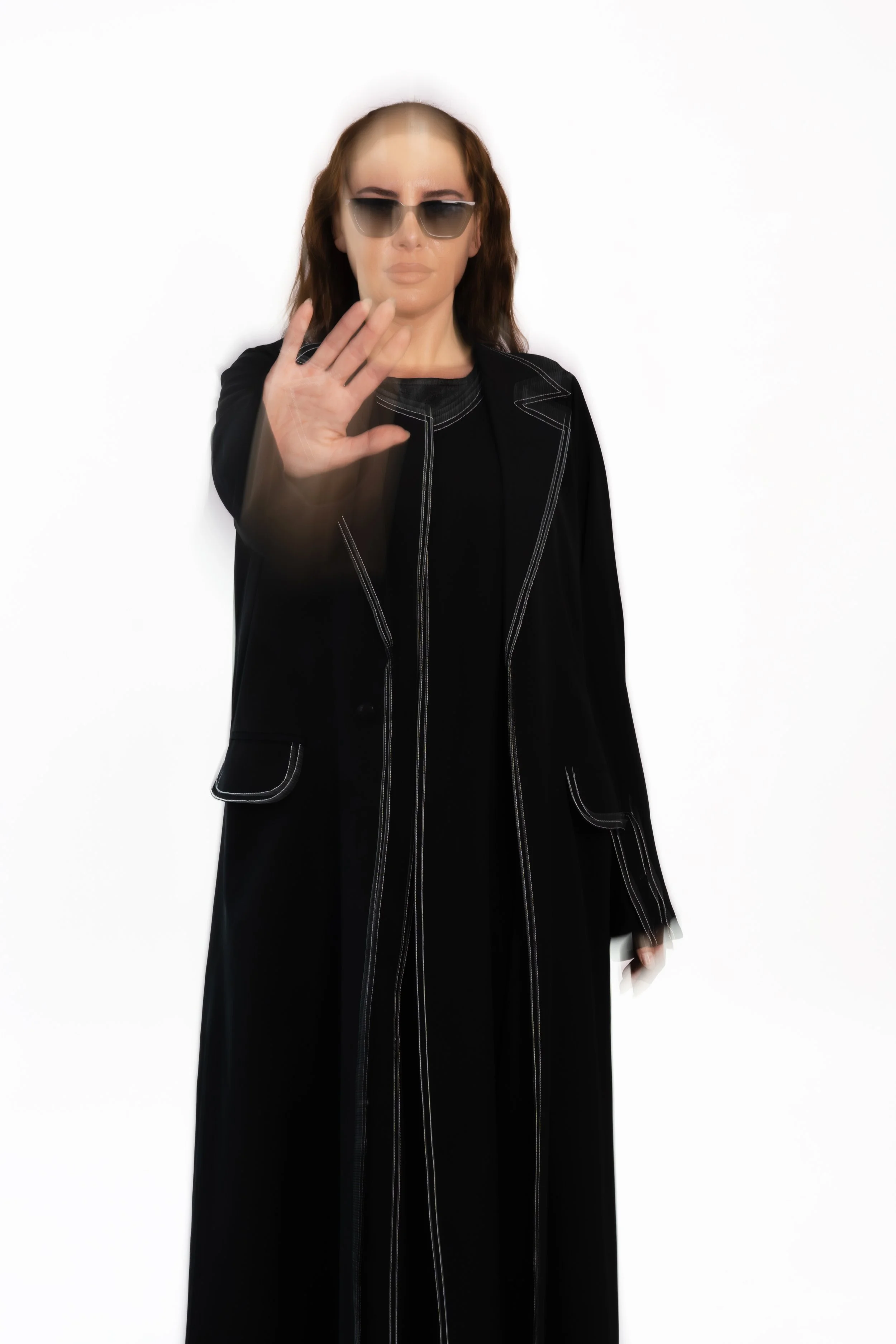 A woman with long brown hair wearing sunglasses and a black long coat with white stitching, extending her hand forward. The background is plain white.