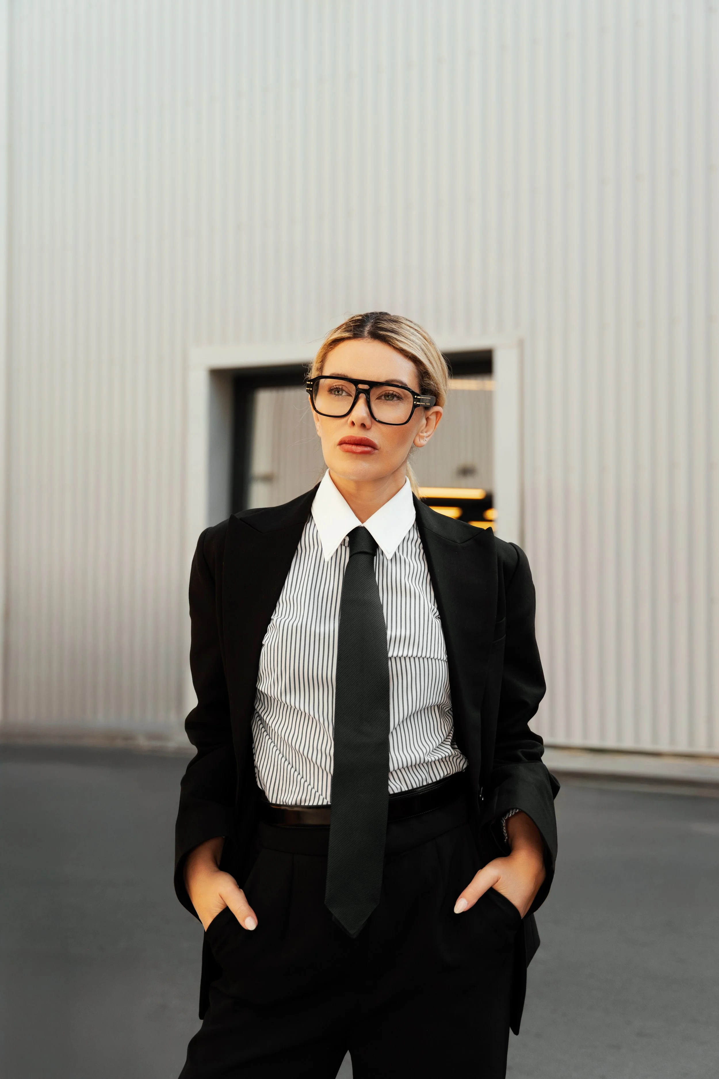 A confident woman stands outside in professional attire, wearing a black blazer, a striped shirt, a black tie, and glasses, with her hands in her pockets and a serious expression.