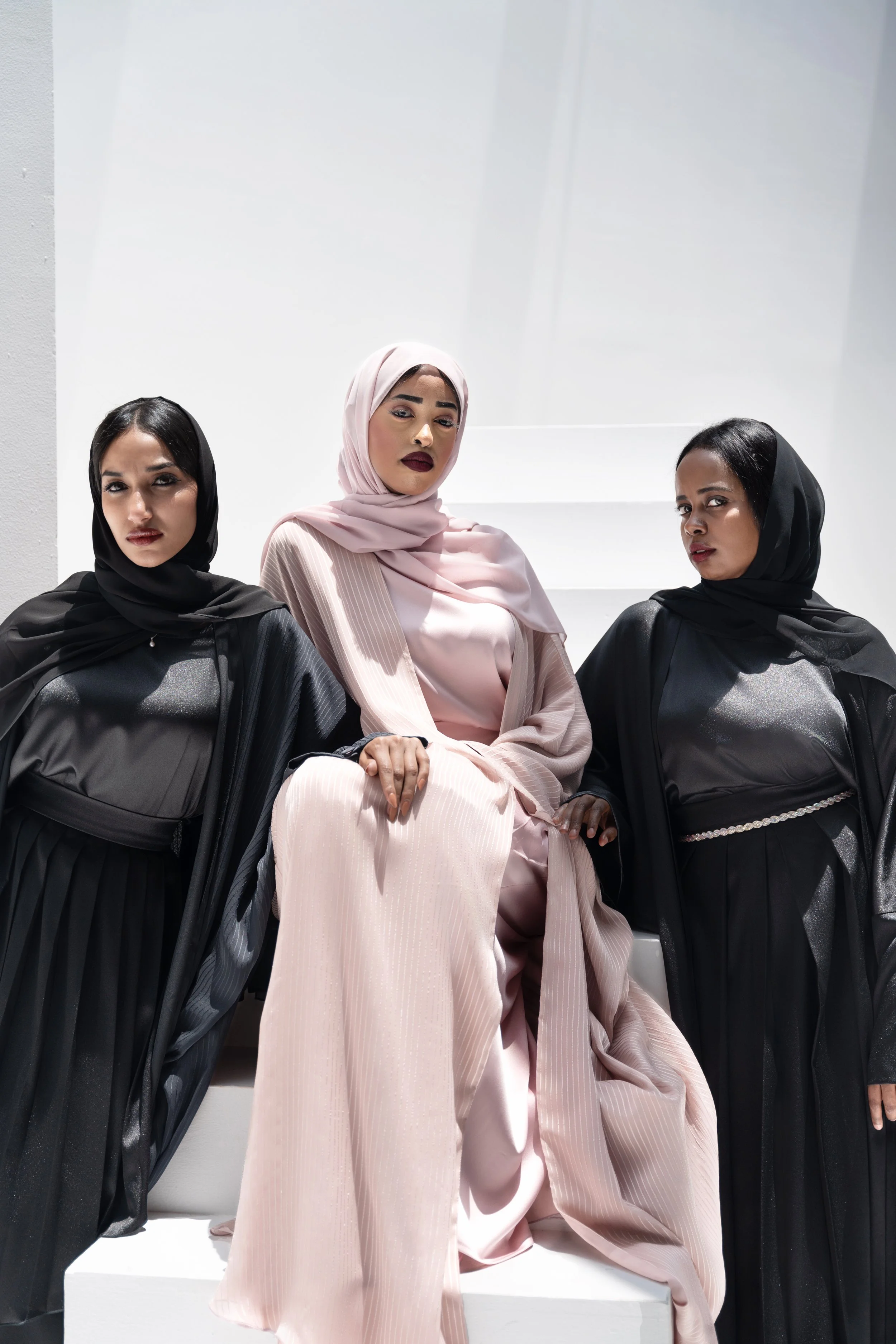 Three women dressed in elegant black and pink outfits and headscarves posing against a white background.