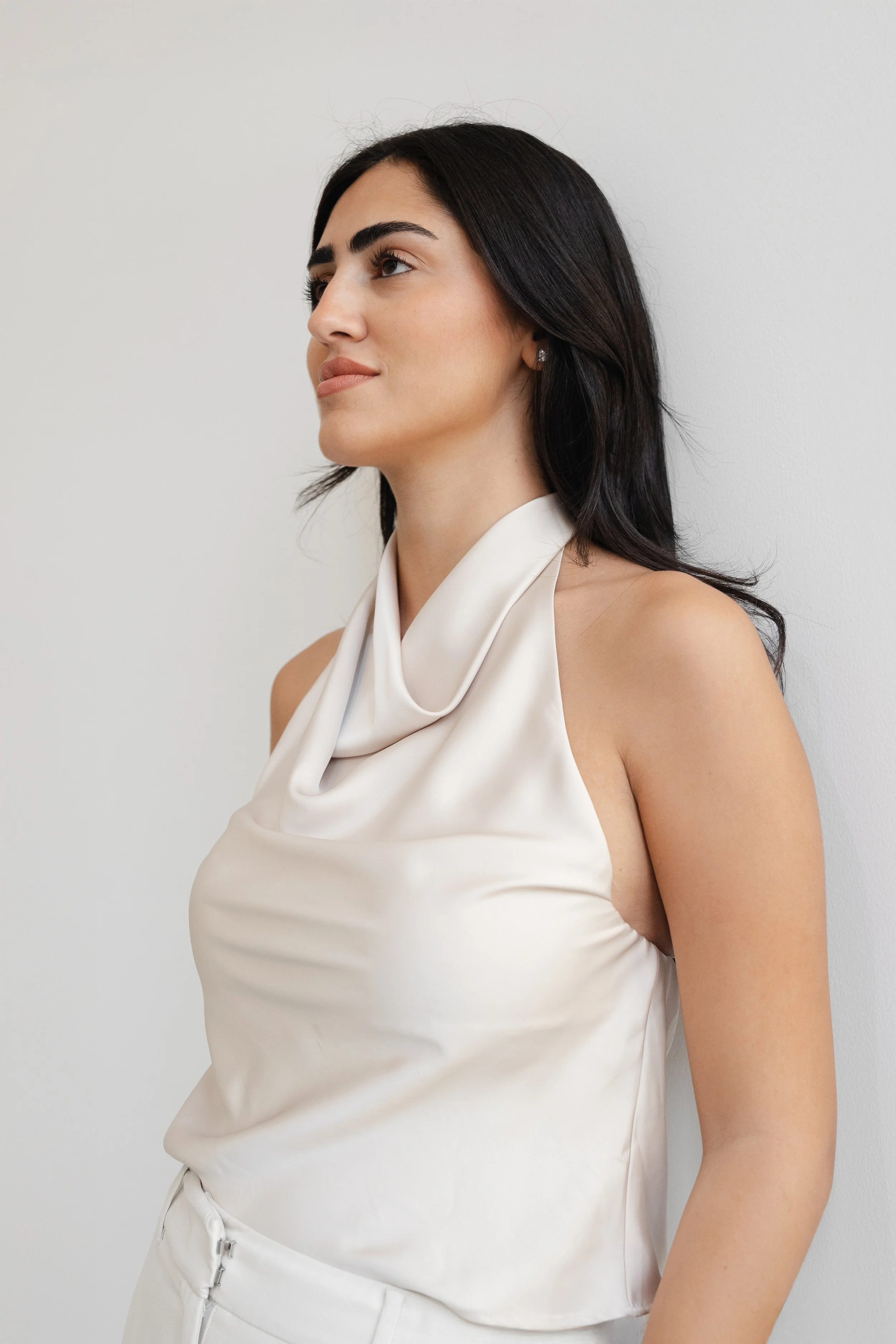 A woman with dark hair, wearing a sleeveless white satin top, stands against a plain white wall.