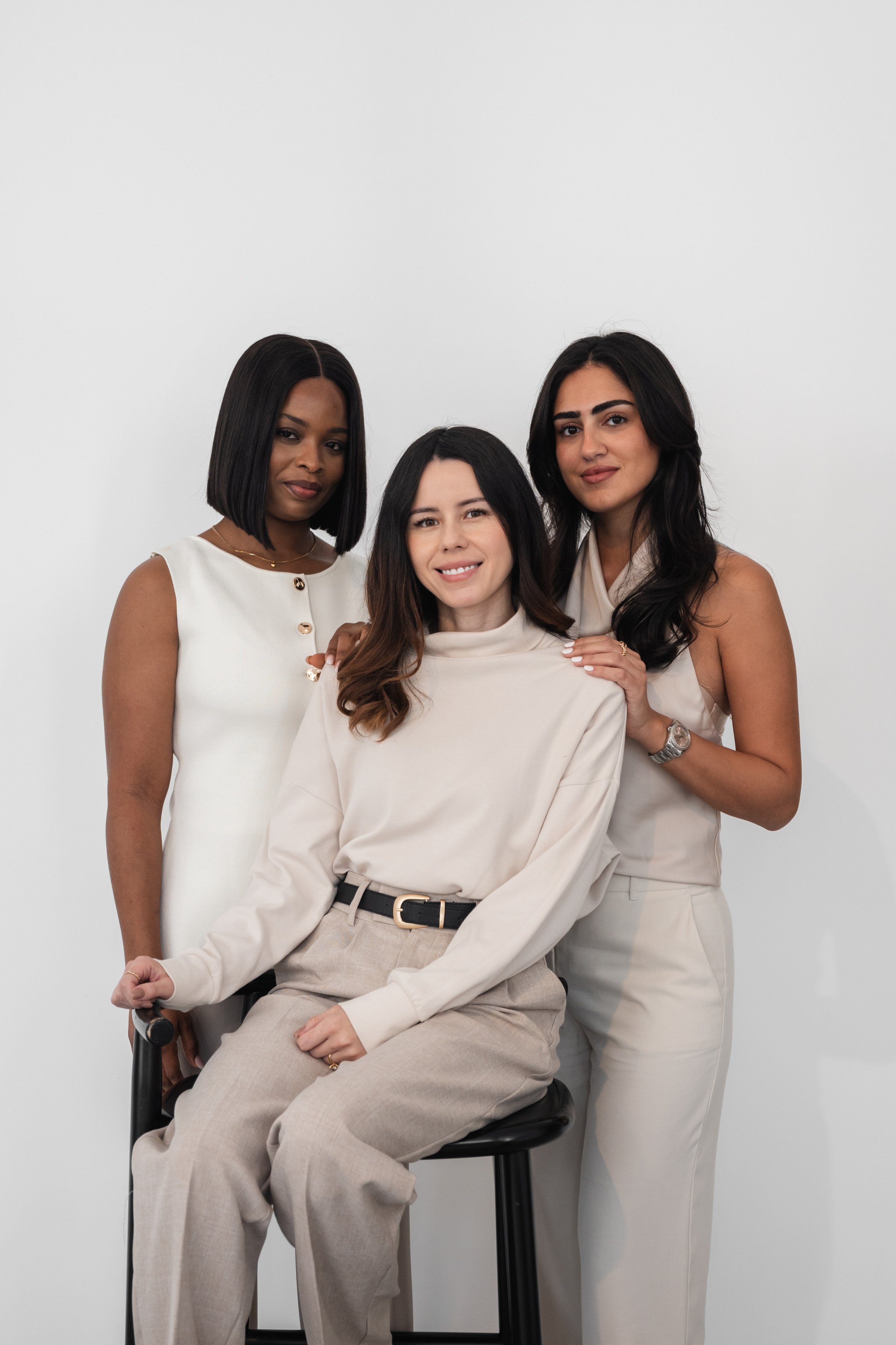 Three women dressed in neutral tones, standing and sitting against a plain white background, posing for a photo.