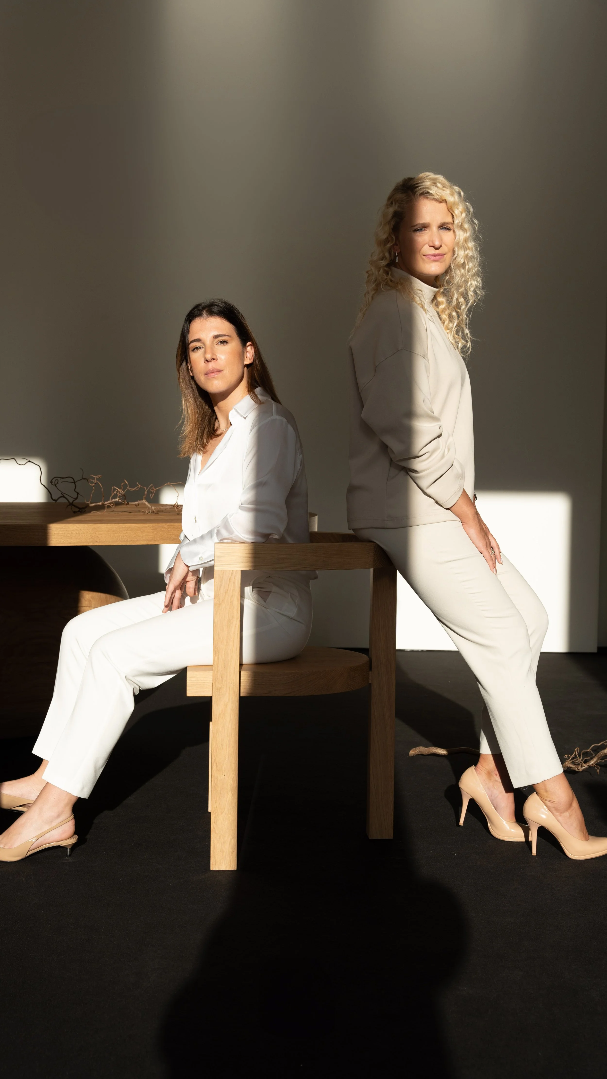 Two women dressed in white outfits sitting and standing in a minimalist room with natural sunlight and shadows.