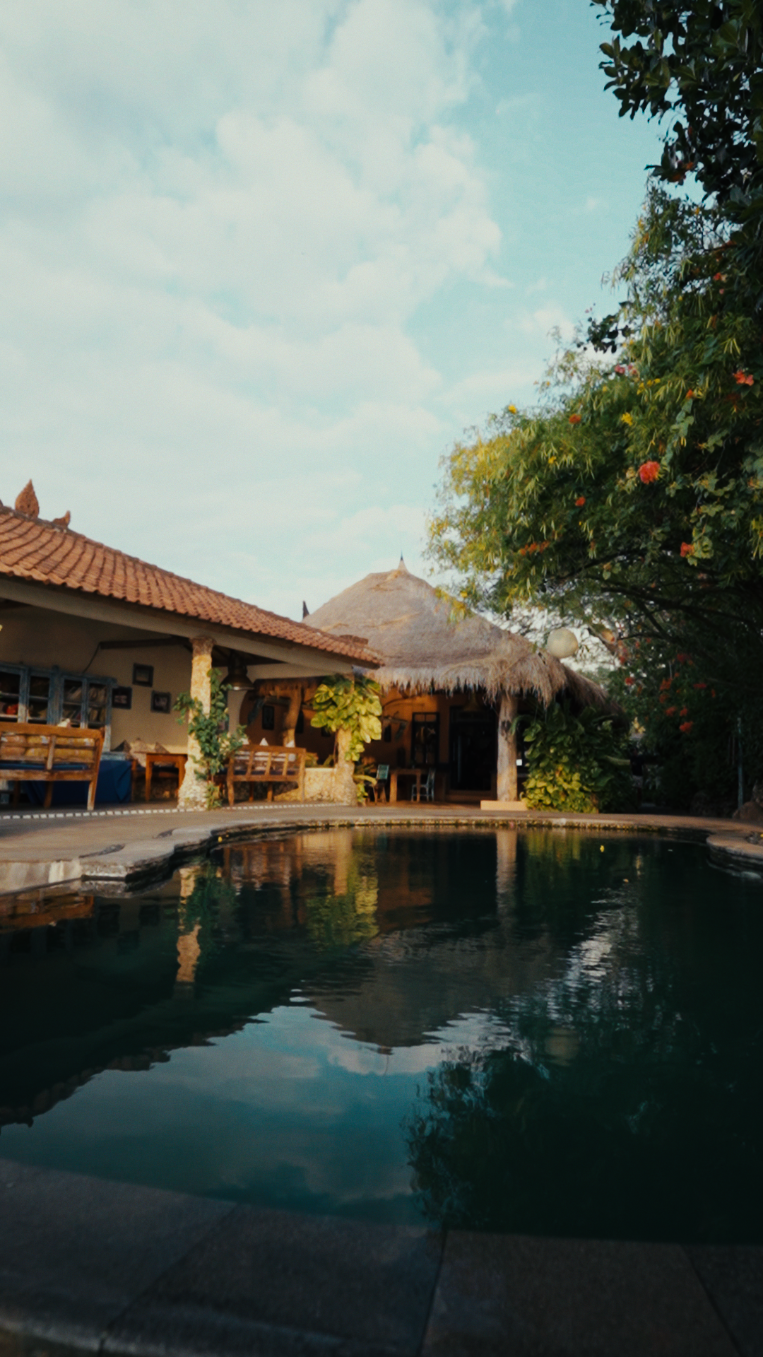 A tropical resort with a swimming pool, thatched roof hut, outdoor seating, lush greenery, and a cloudy sky.
