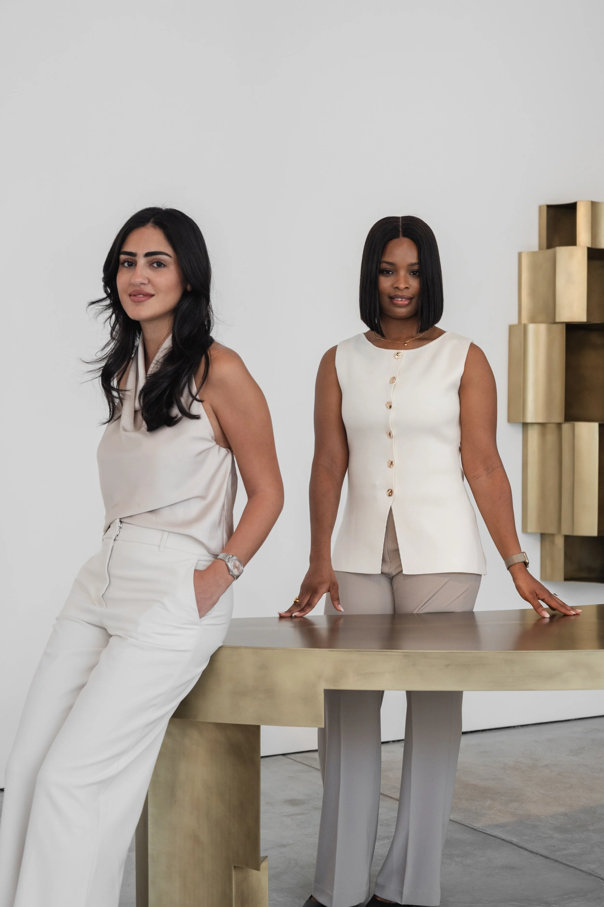 Two women in neutral-colored clothing standing in a modern, minimalistic room with a gold desk and abstract gold wall art.