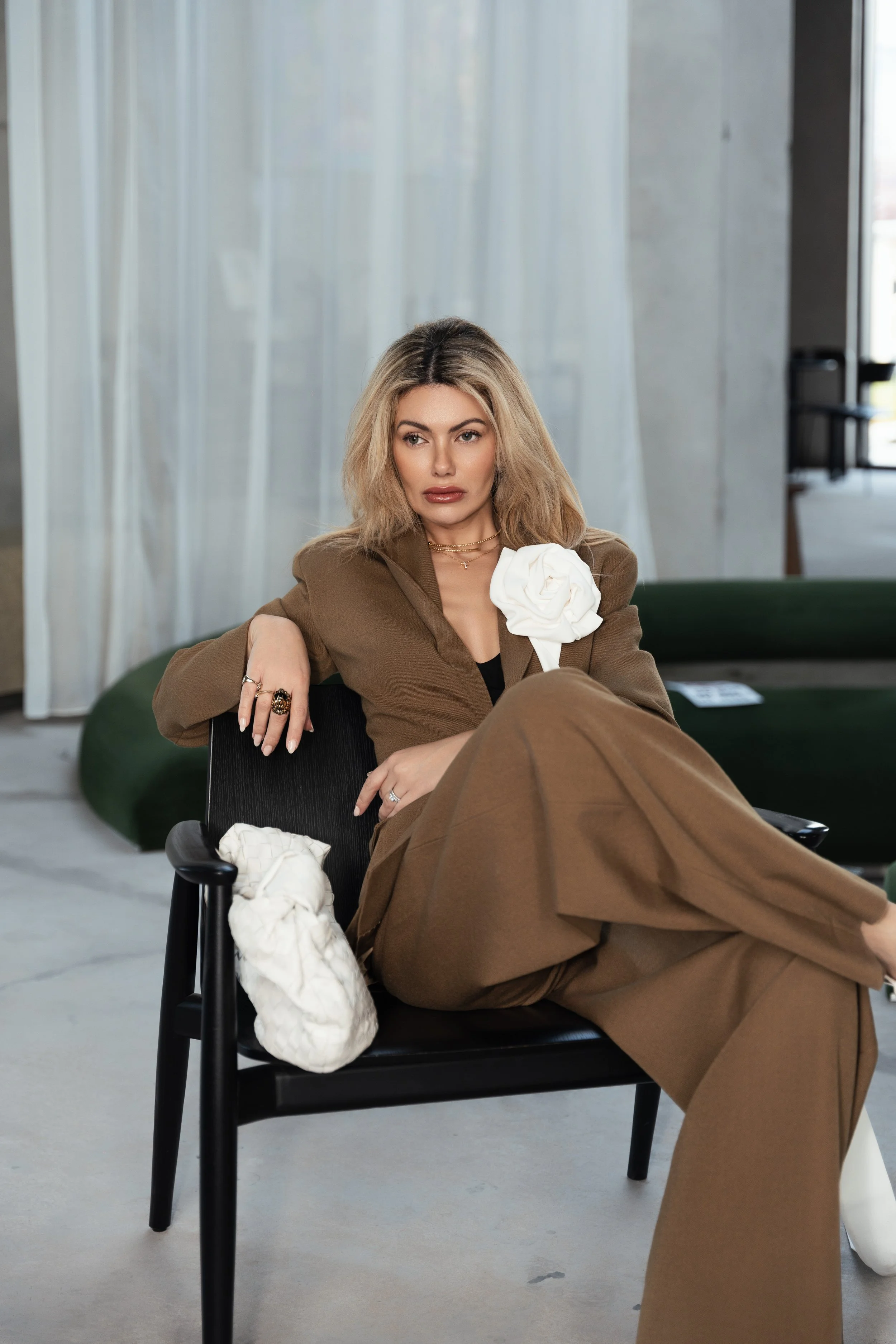 A woman with blonde hair, wearing brown pants and blazer, sitting on a black chair in a modern interior space with white curtains and green furniture.