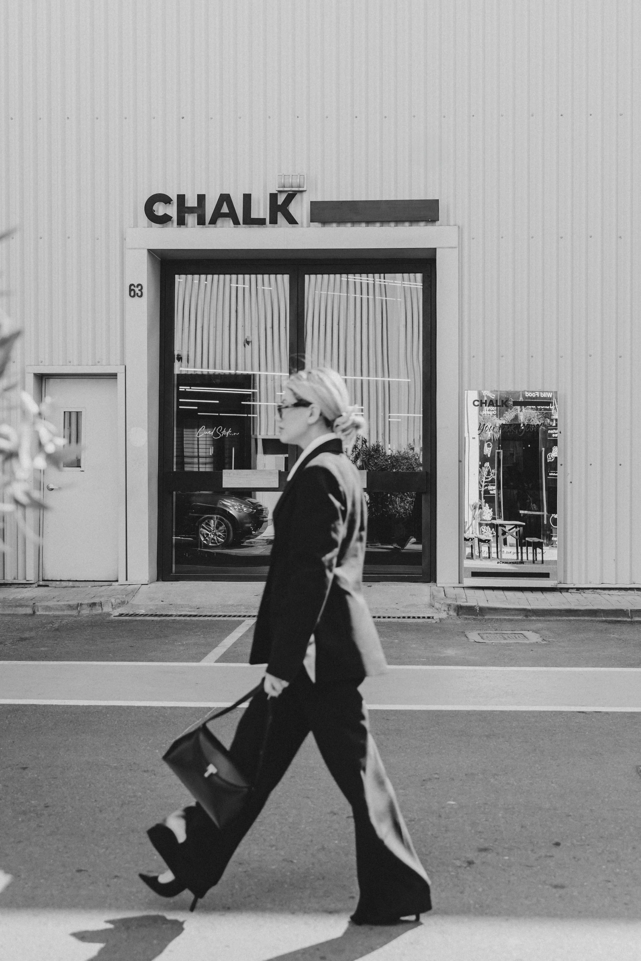 A woman with blonde hair, sunglasses, and a black suit walking across the street carrying a black bag in front of a business called CHALK.