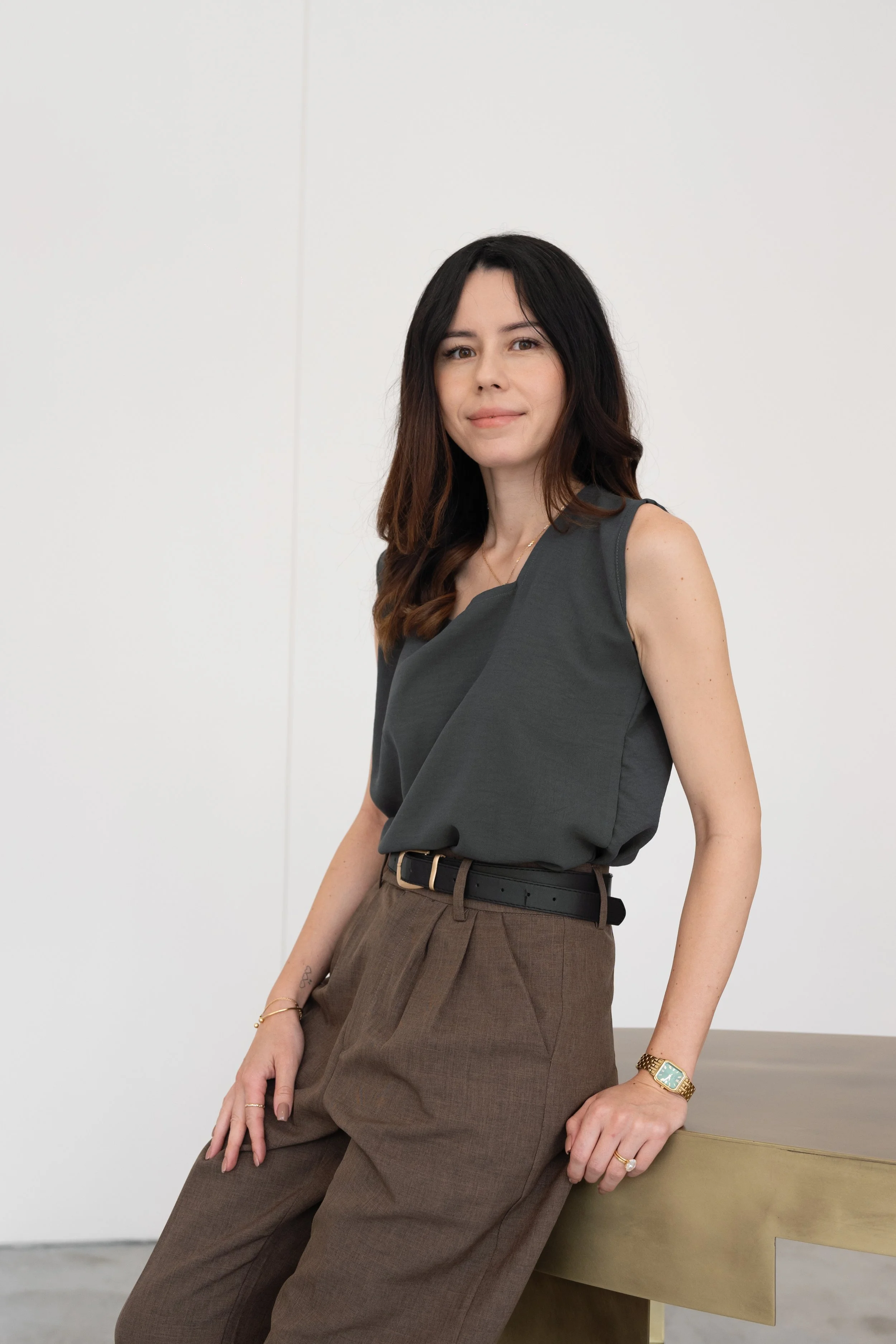 A woman with long dark hair wearing a dark sleeveless top and brown trousers, sitting on a wooden table against a plain white background.