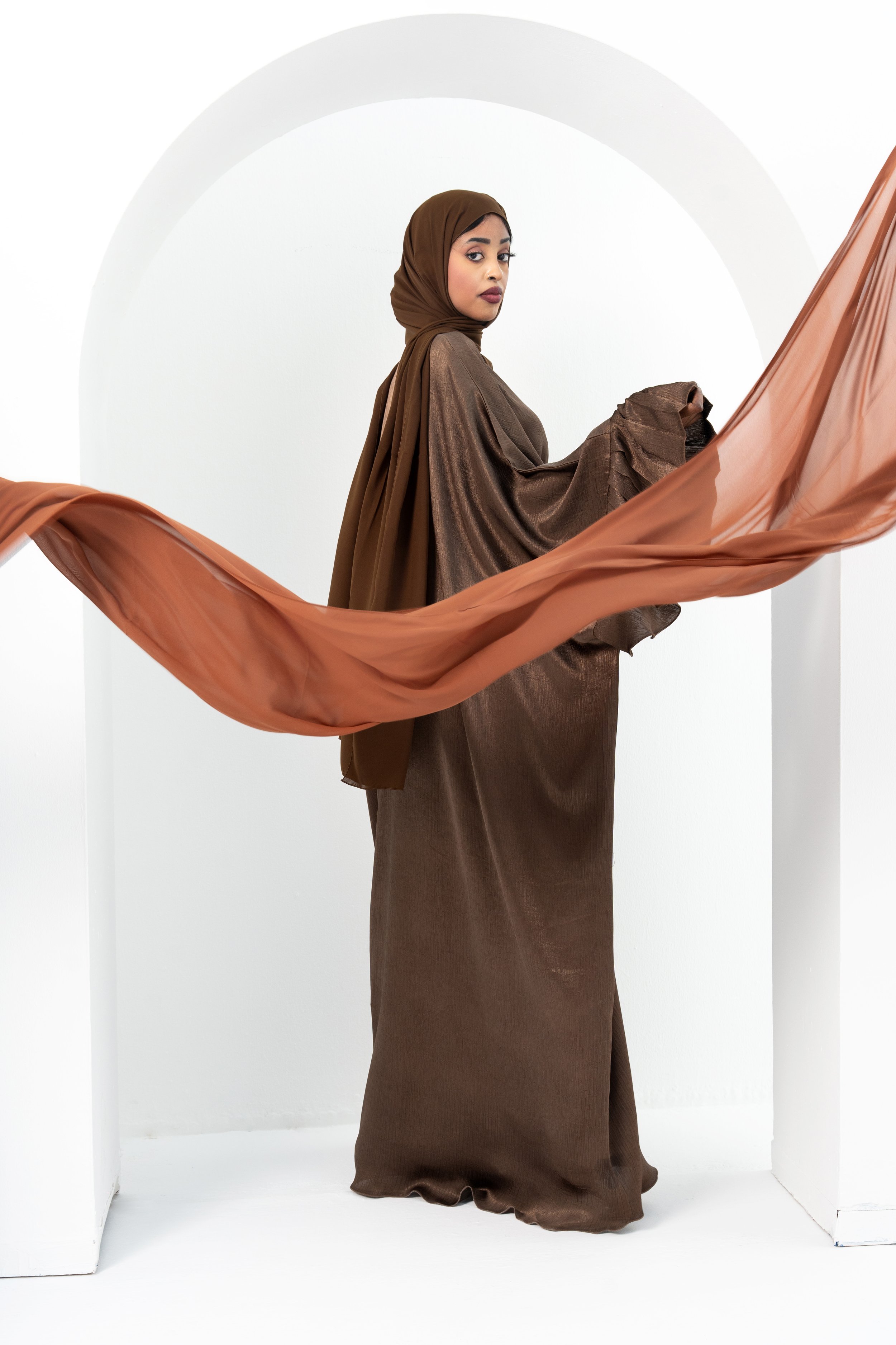 A woman wearing a brown hijab and flowy brown dress stands in a white minimalist studio with an arched backdrop, holding an orange fabric that extends outward.