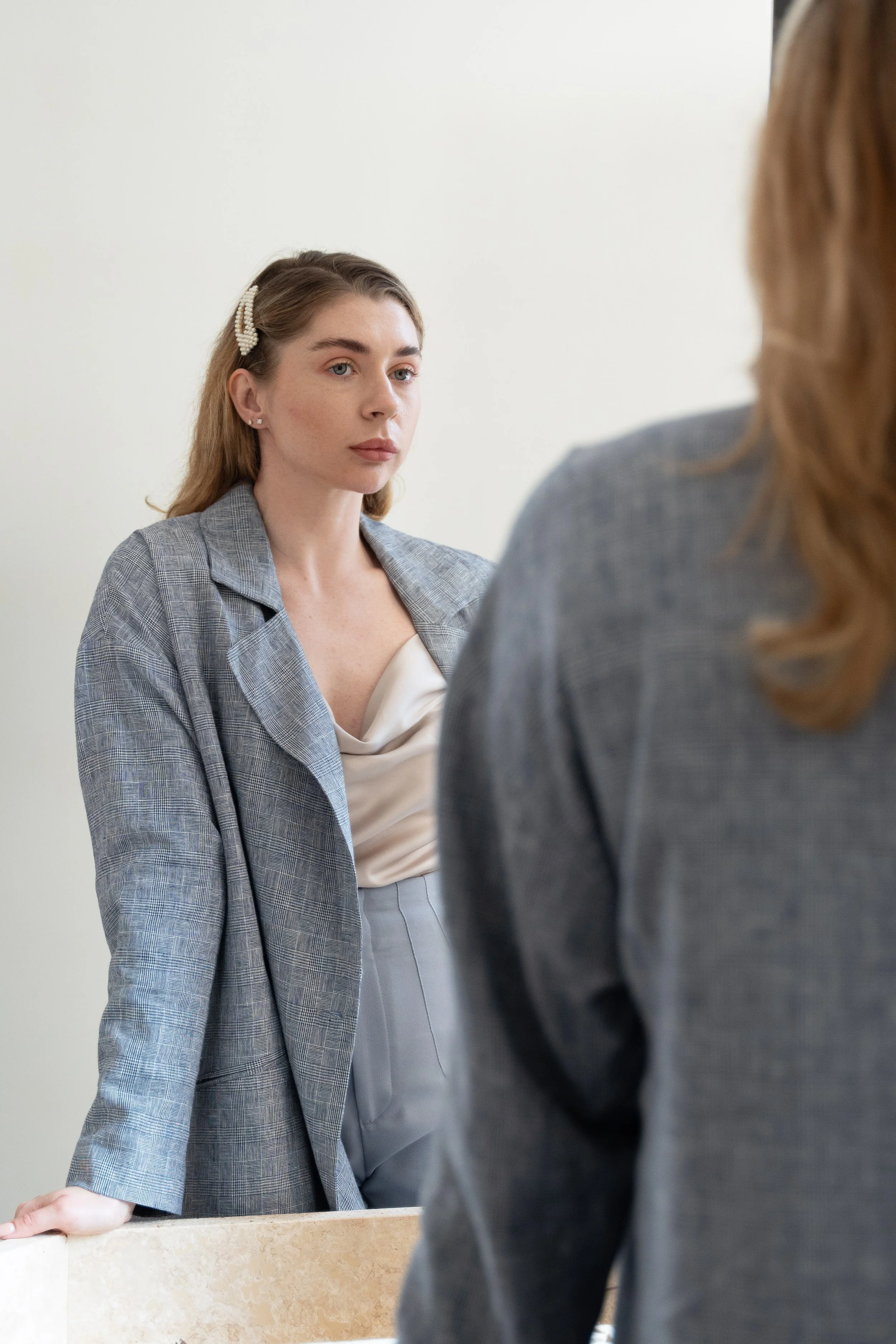 Woman with blonde hair wearing a gray blazer and a cream satin top, standing quietly and looking thoughtfully at another figure in a similar gray blazer, with a plain light-colored wall background.