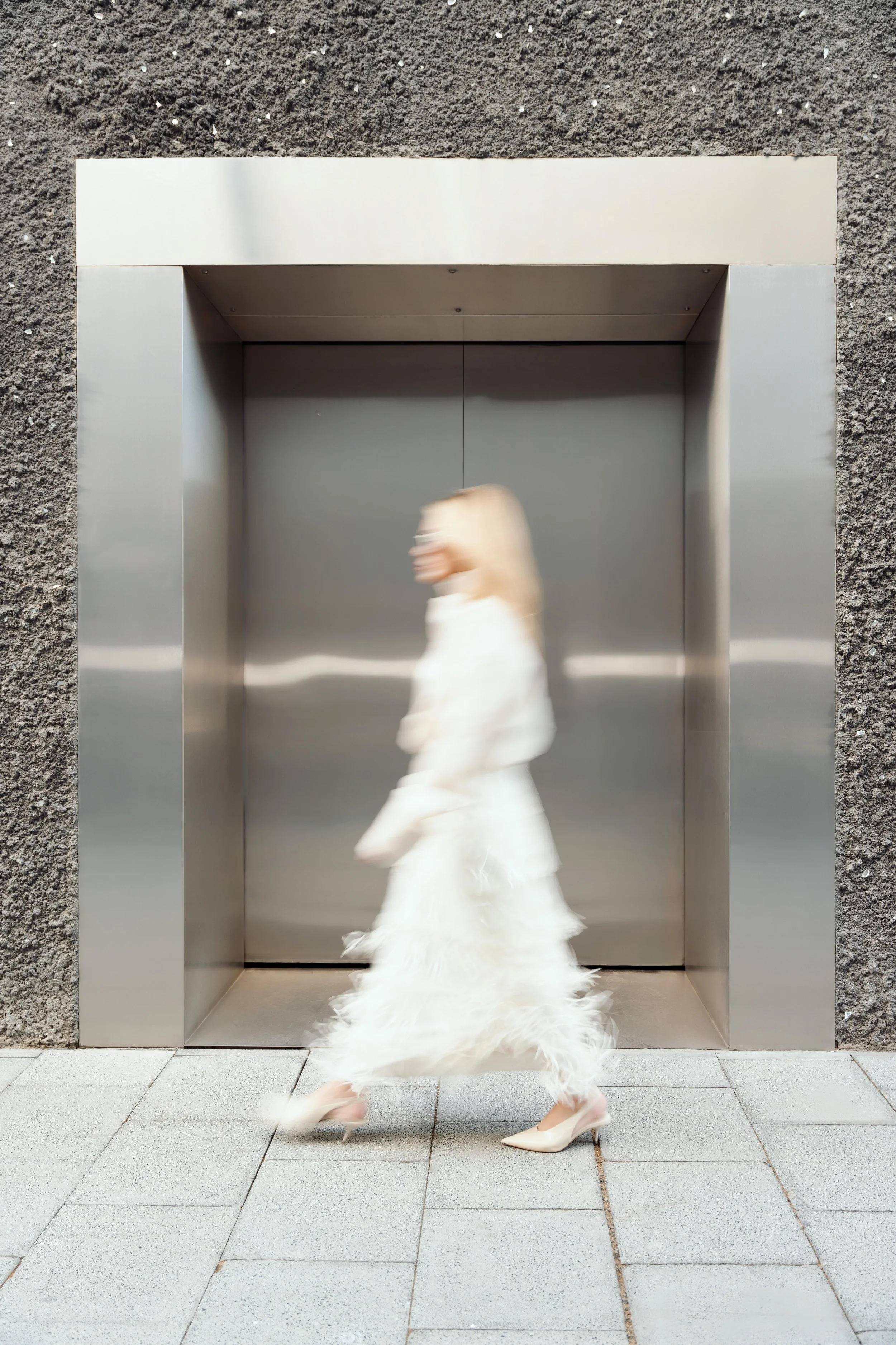 A woman walking past a modern elevator door with a textured stone wall and tiled sidewalk in an urban setting