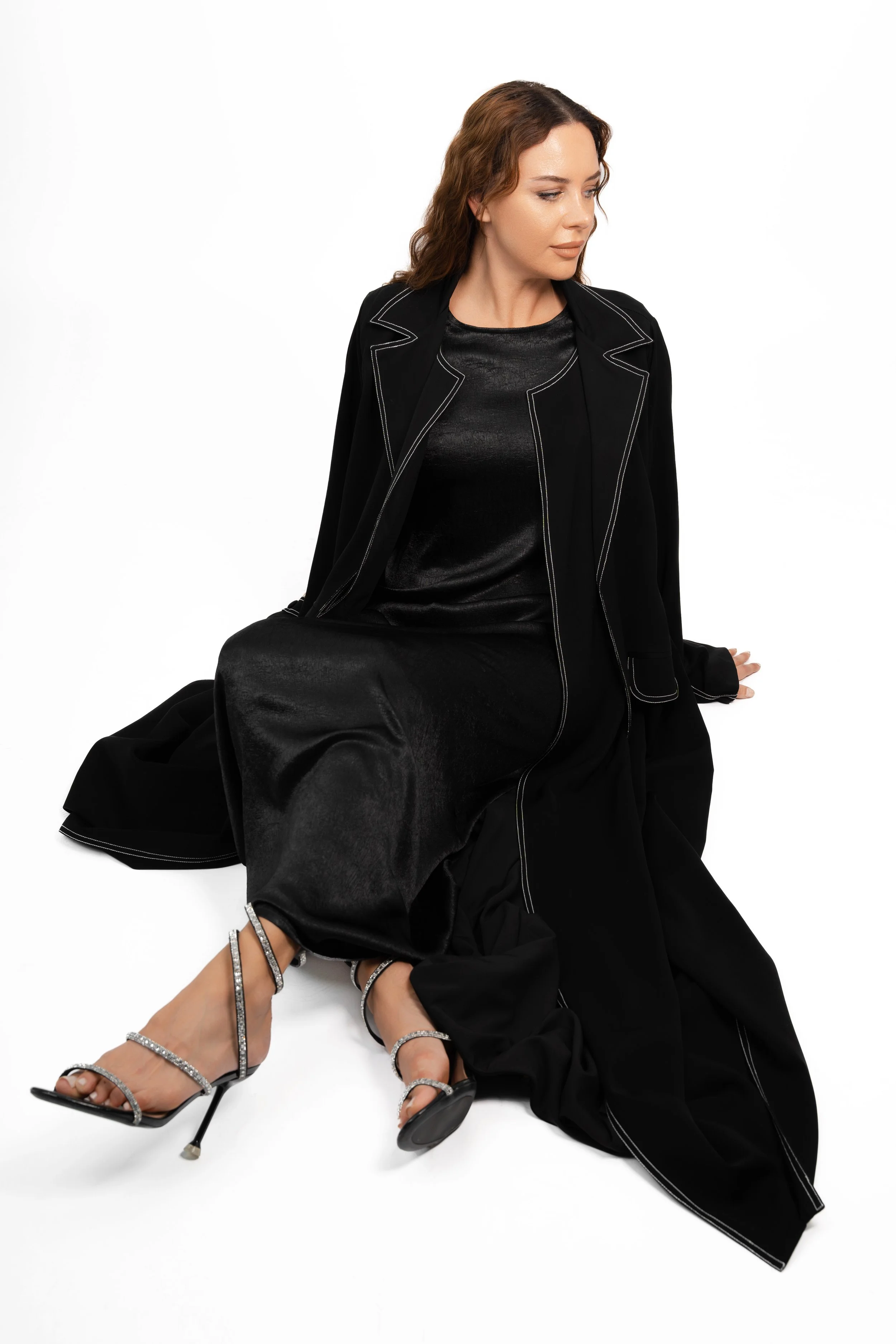 A woman wearing a black satin dress, black coat with white stitching, and silver high-heeled sandals with rhinestones, sitting on the floor against a white background.