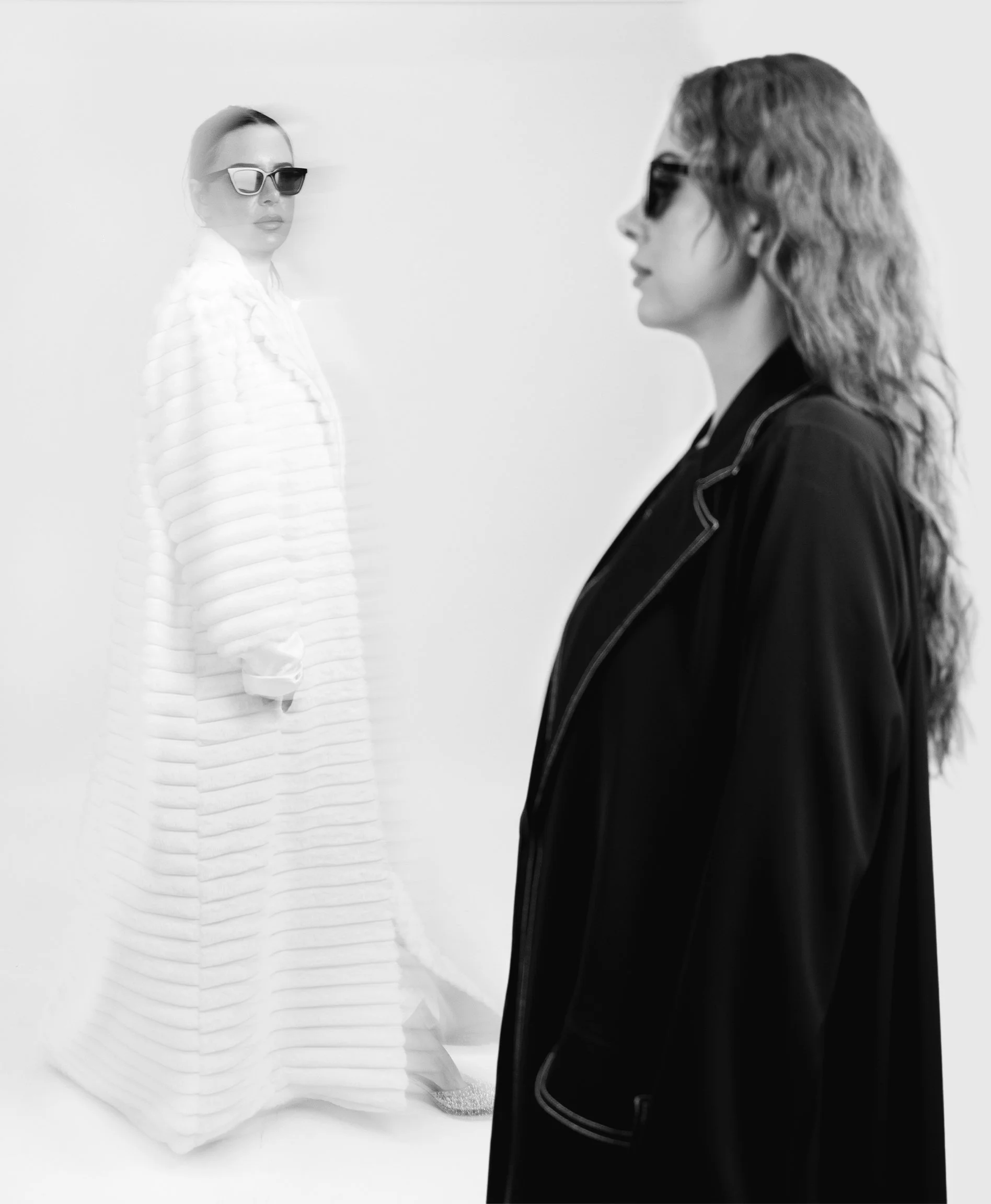 Two women wearing sunglasses and dark clothing, standing apart in an indoor photo, in a black and white style.