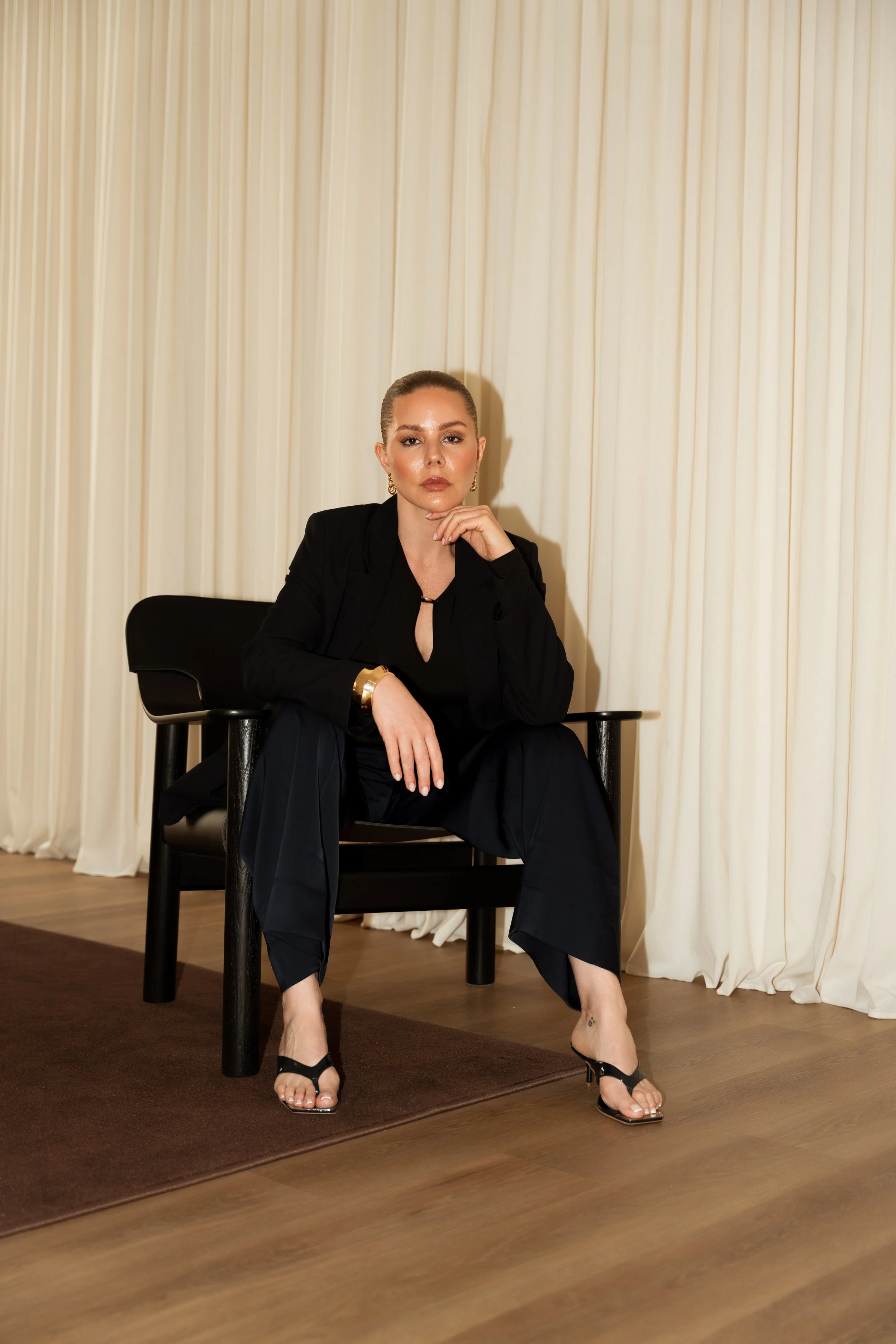 A woman sitting on a black chair in front of cream-colored curtains, dressed in a black blazer, black top, black pants, and black high-heeled sandals, with her leg crossed and her hand resting near her chin