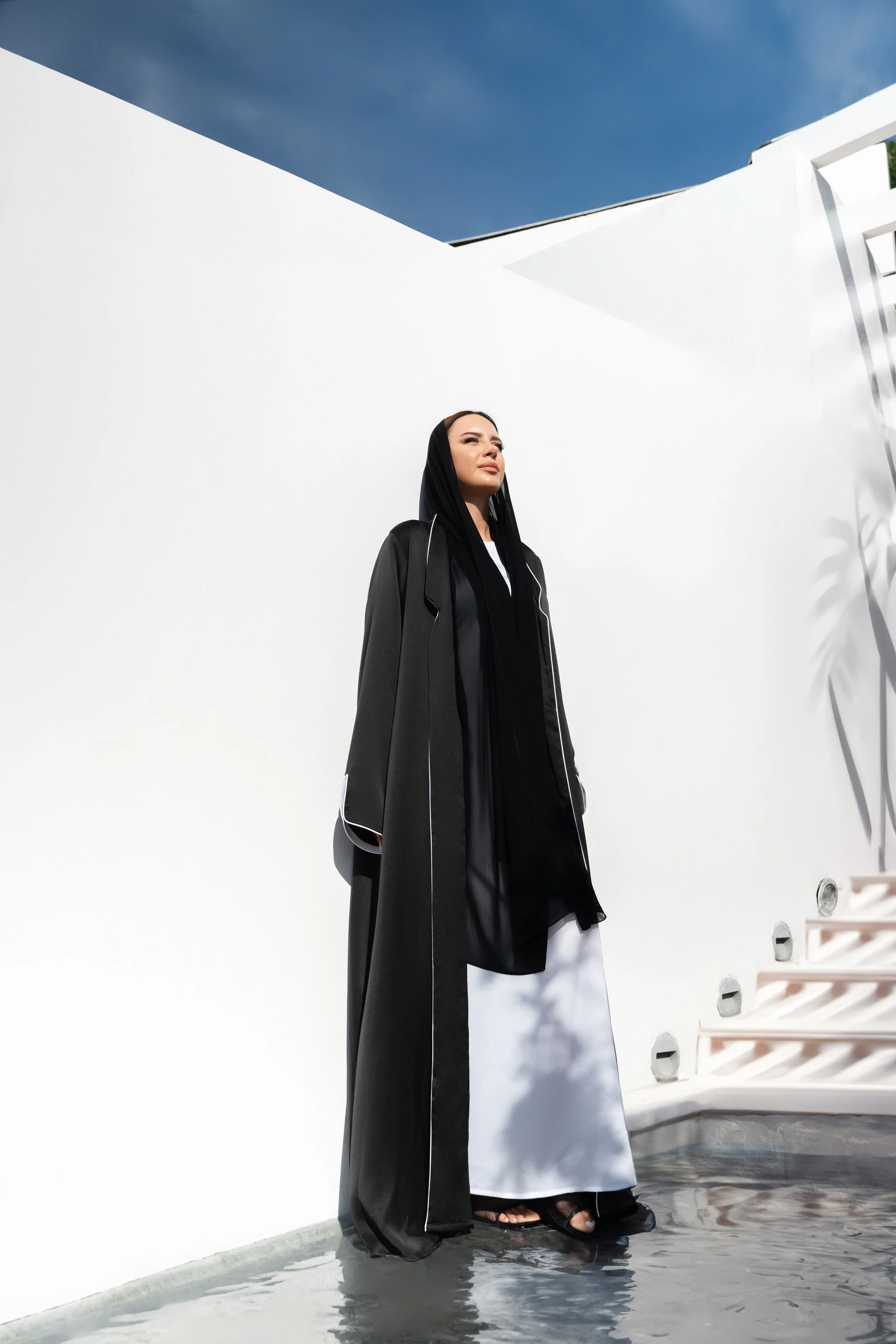 A woman wearing a black and white abaya stands on a reflective surface outdoors against a white wall with blue sky and clouds above.