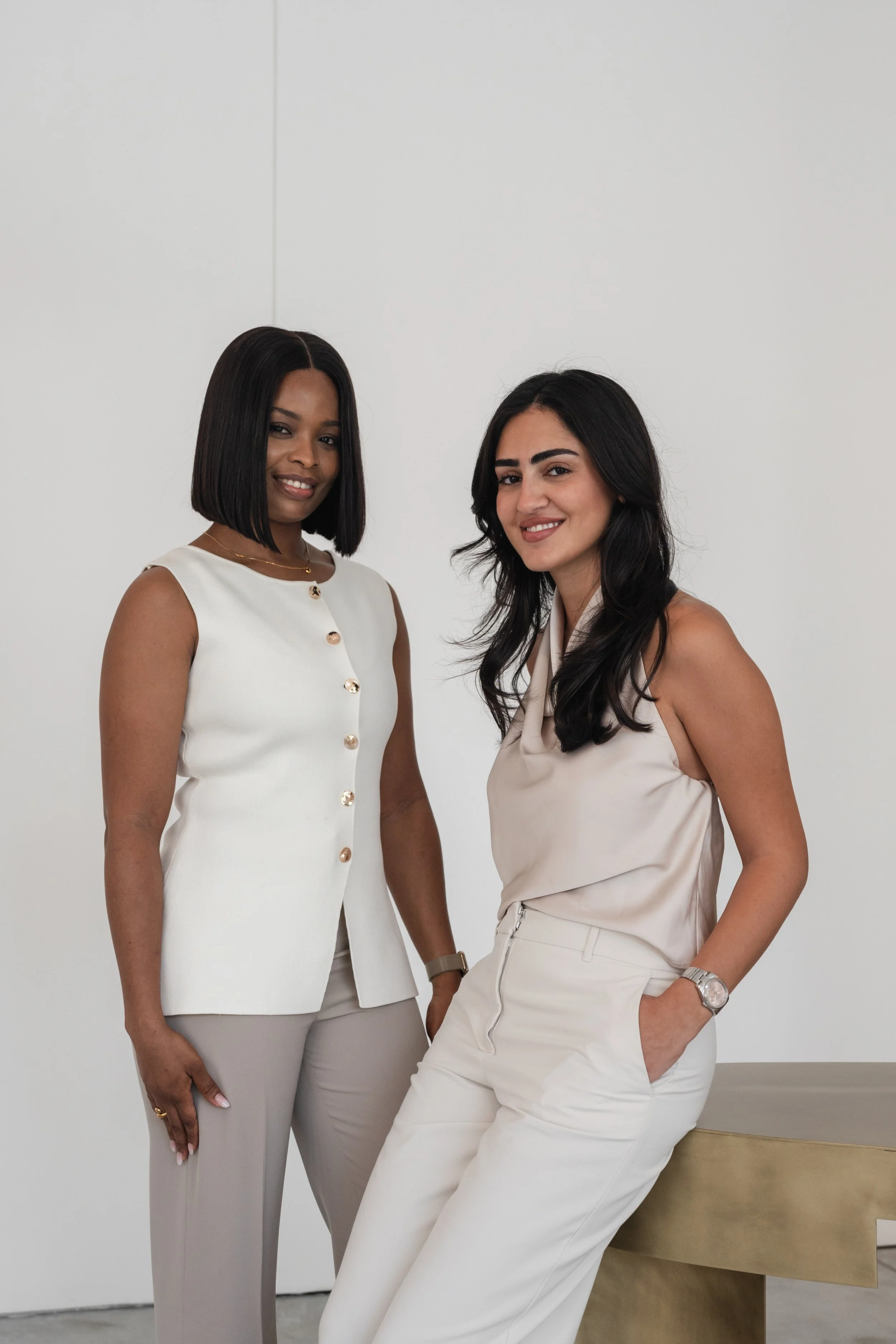 Two women in professional attire standing against a plain white wall, smiling at the camera.
