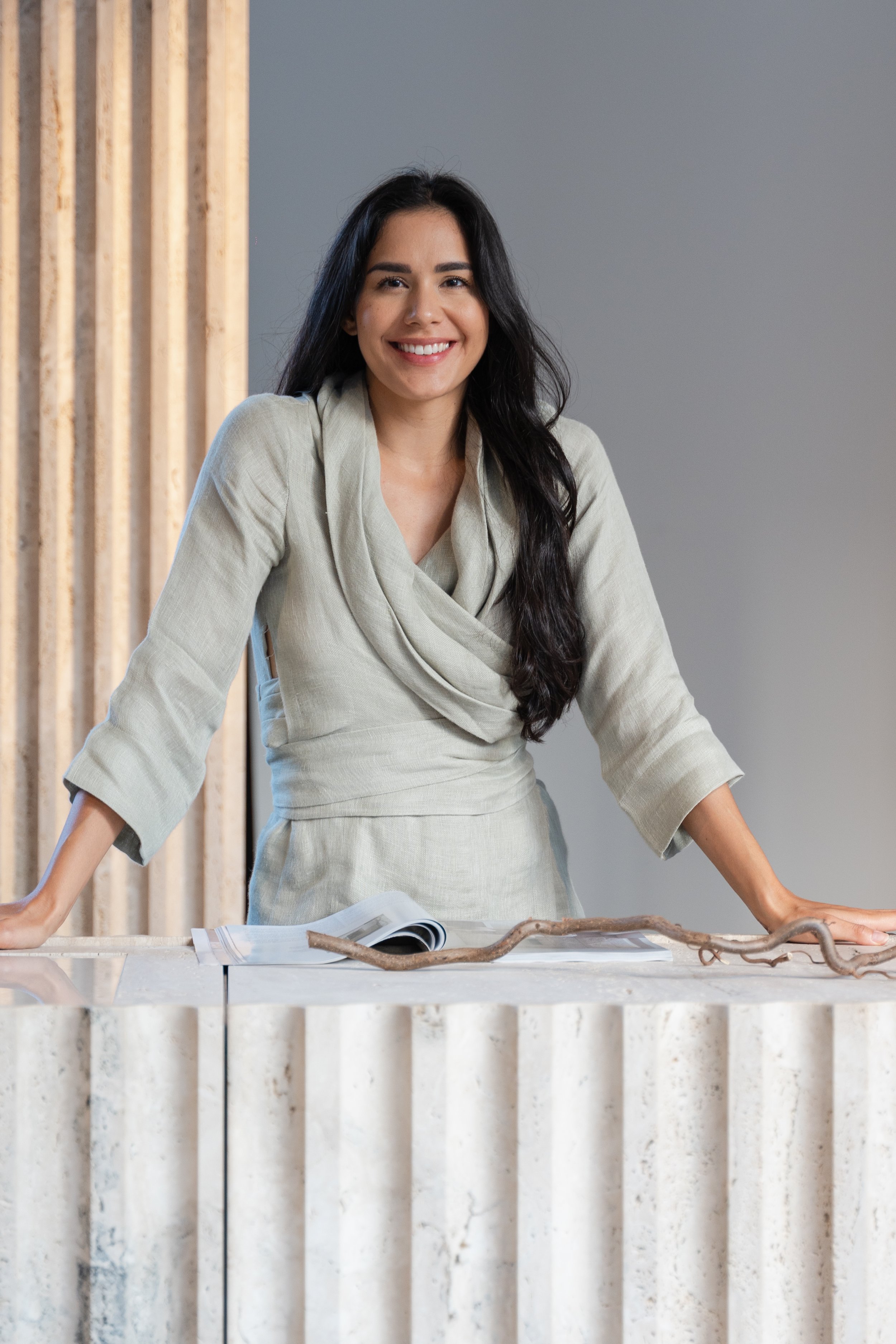 A woman with long dark hair smiling and leaning on a marble counter with an open magazine and a decorative branch on it, standing in front of a modern interior backdrop.