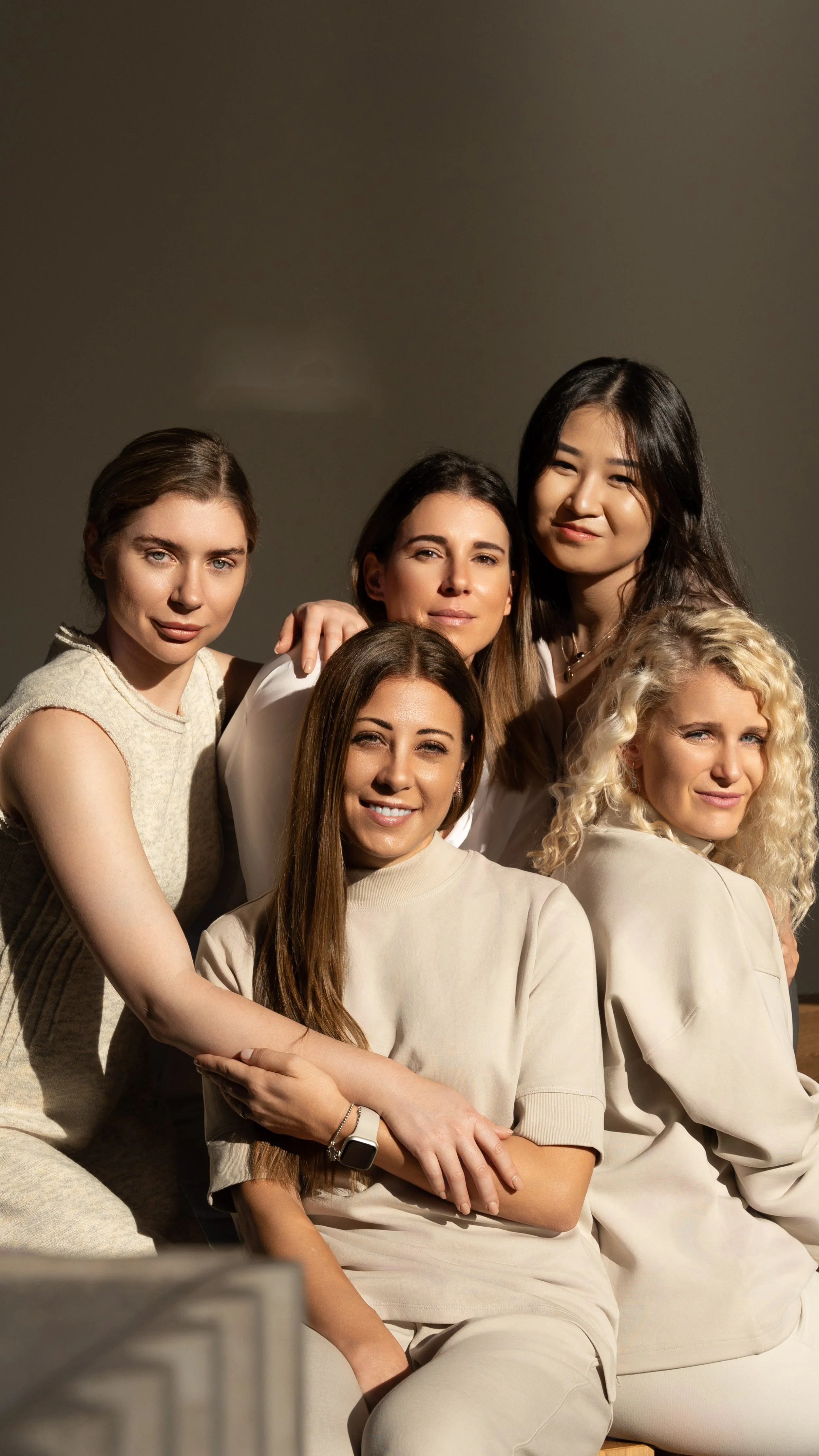 A group of five women seated close together, smiling at the camera, in a studio with a plain background.