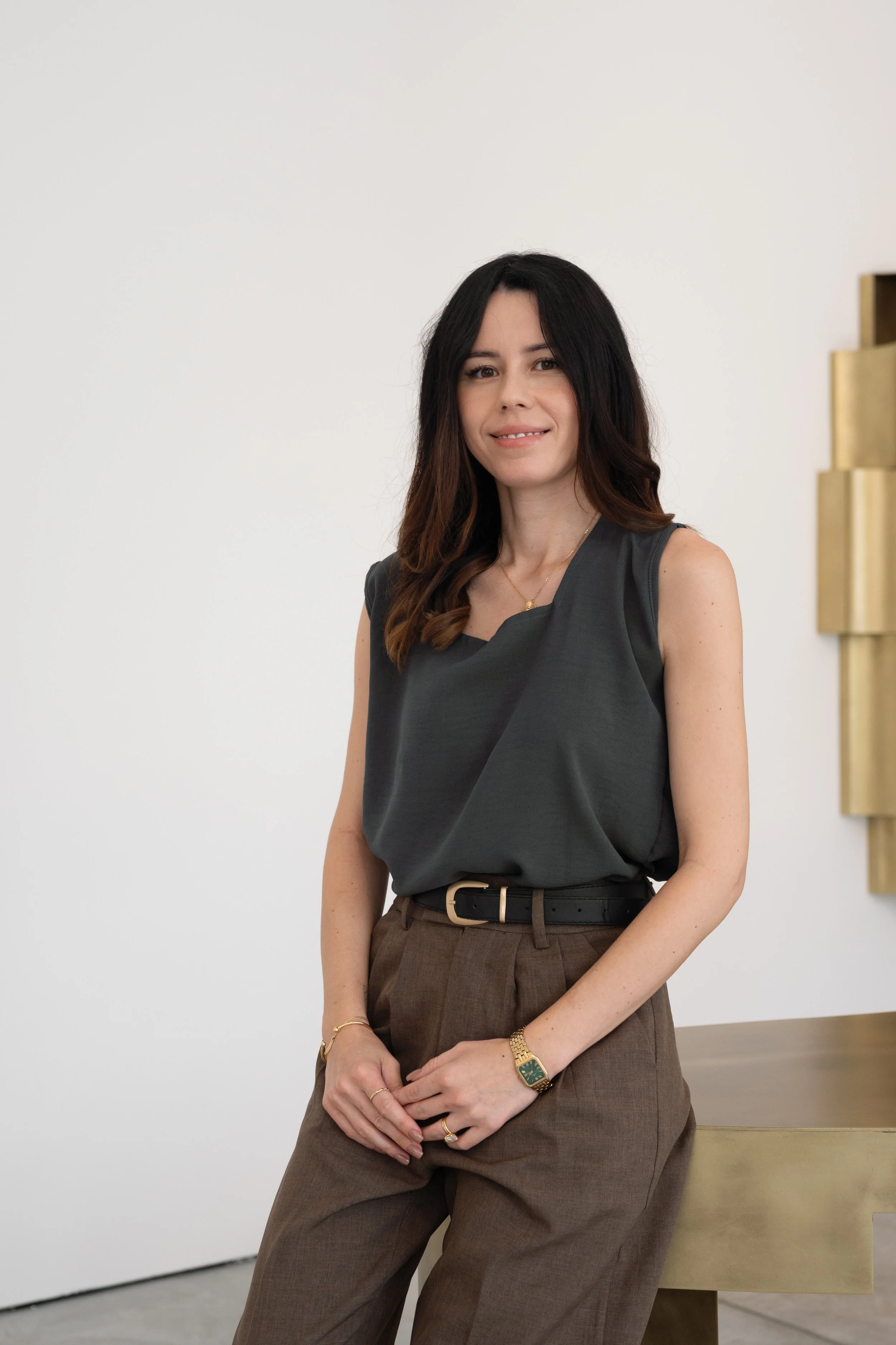 A woman with long dark hair, wearing a dark sleeveless blouse and brown trousers, standing in front of a white wall with gold decorative elements on the right side.
