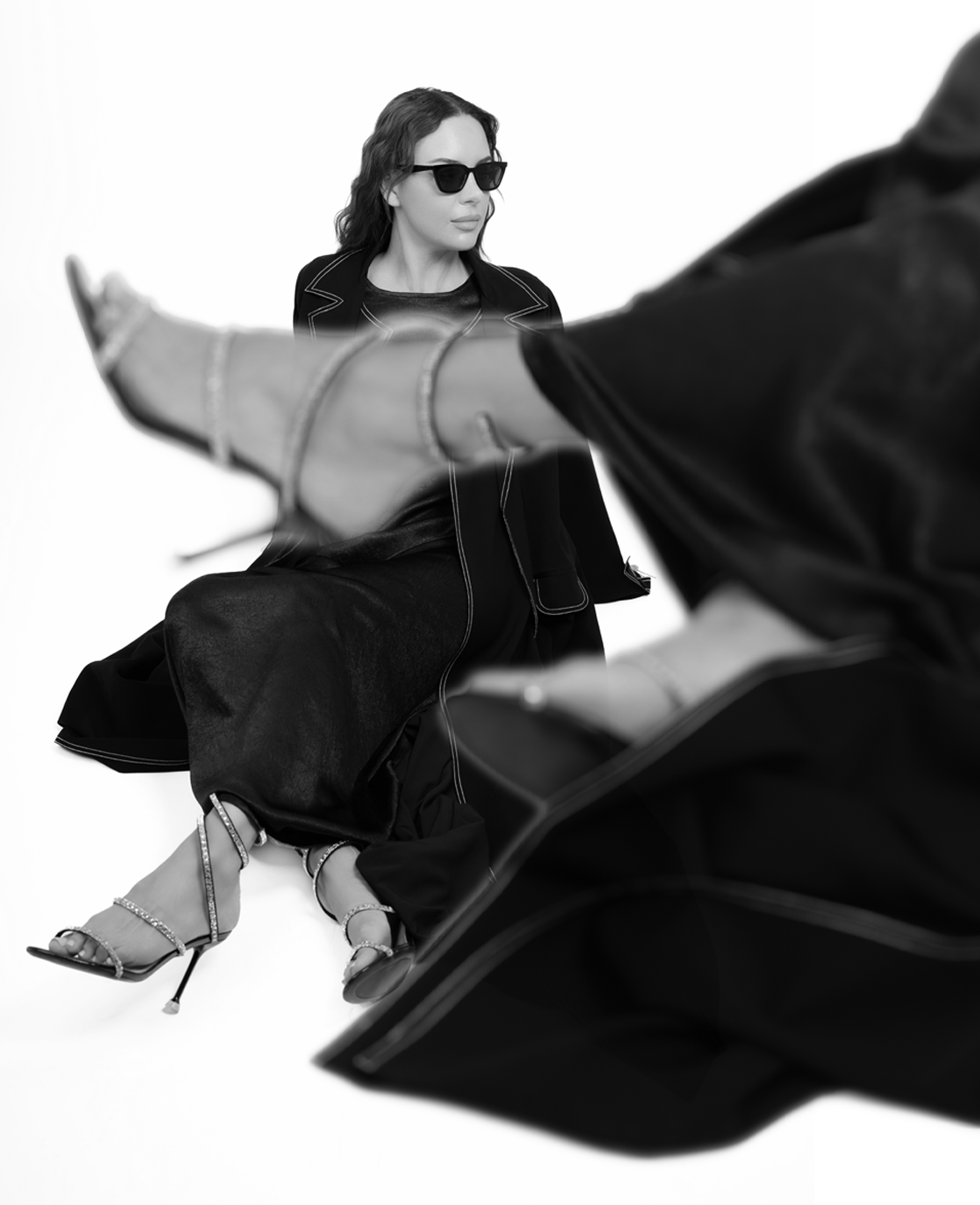 Black and white photo of a woman wearing sunglasses and a dark outfit, sitting with one leg crossed over the other while a person in the foreground reaches out with an open hand.