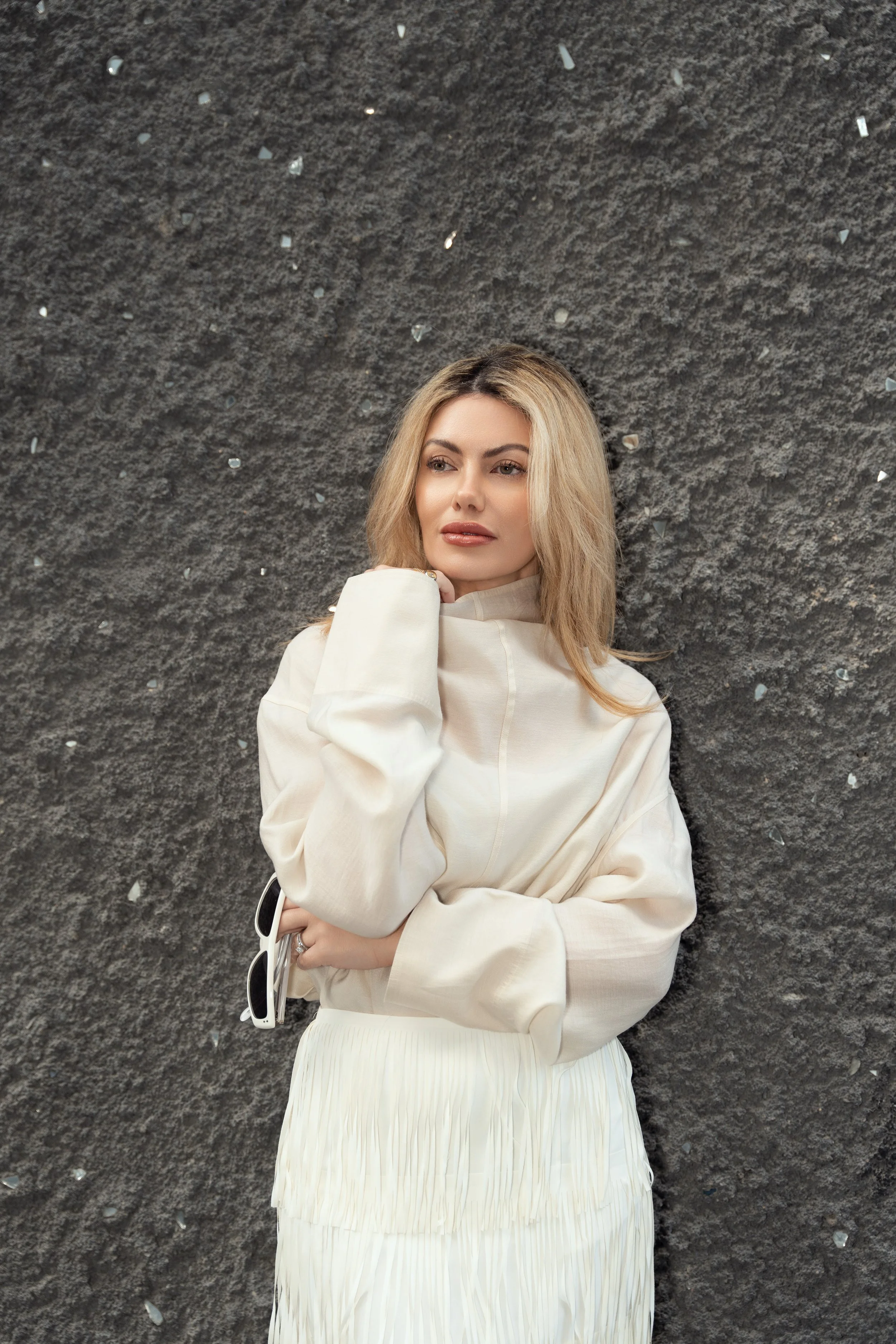 A woman with blonde hair in a beige top and white skirt leaning against a textured dark gray wall.