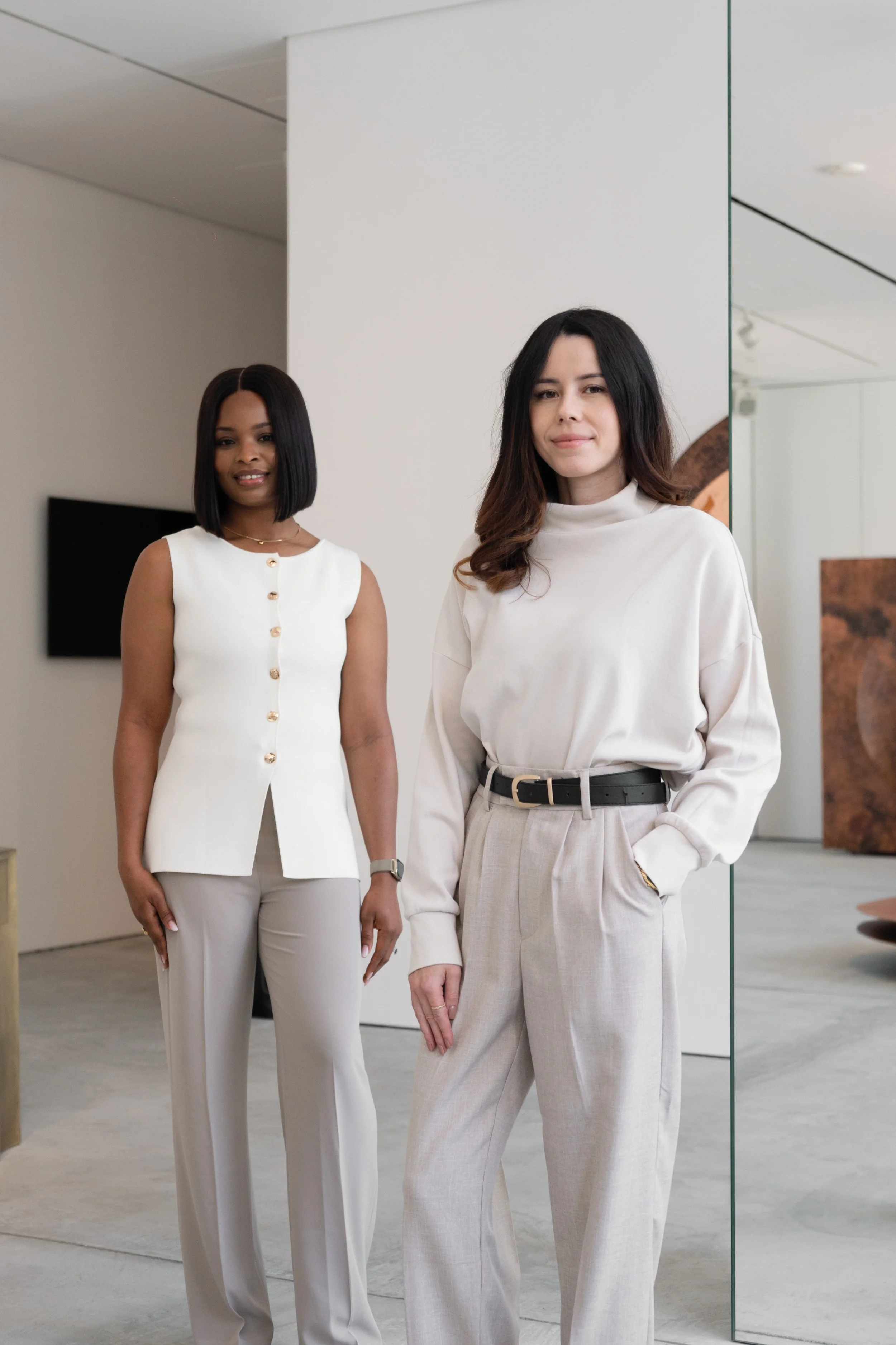 Two women standing indoors, both dressed in neutral-colored business casual outfits, posing for the photo.