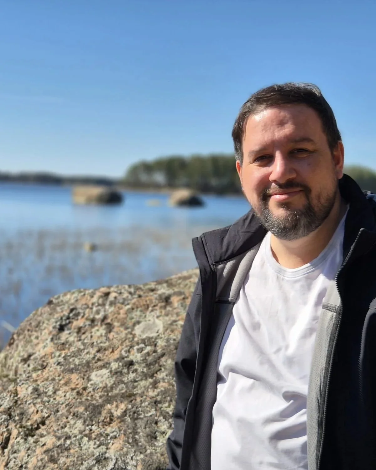 A man with a beard and short hair smiling outdoors near a body of water with rocks and trees in the background.
